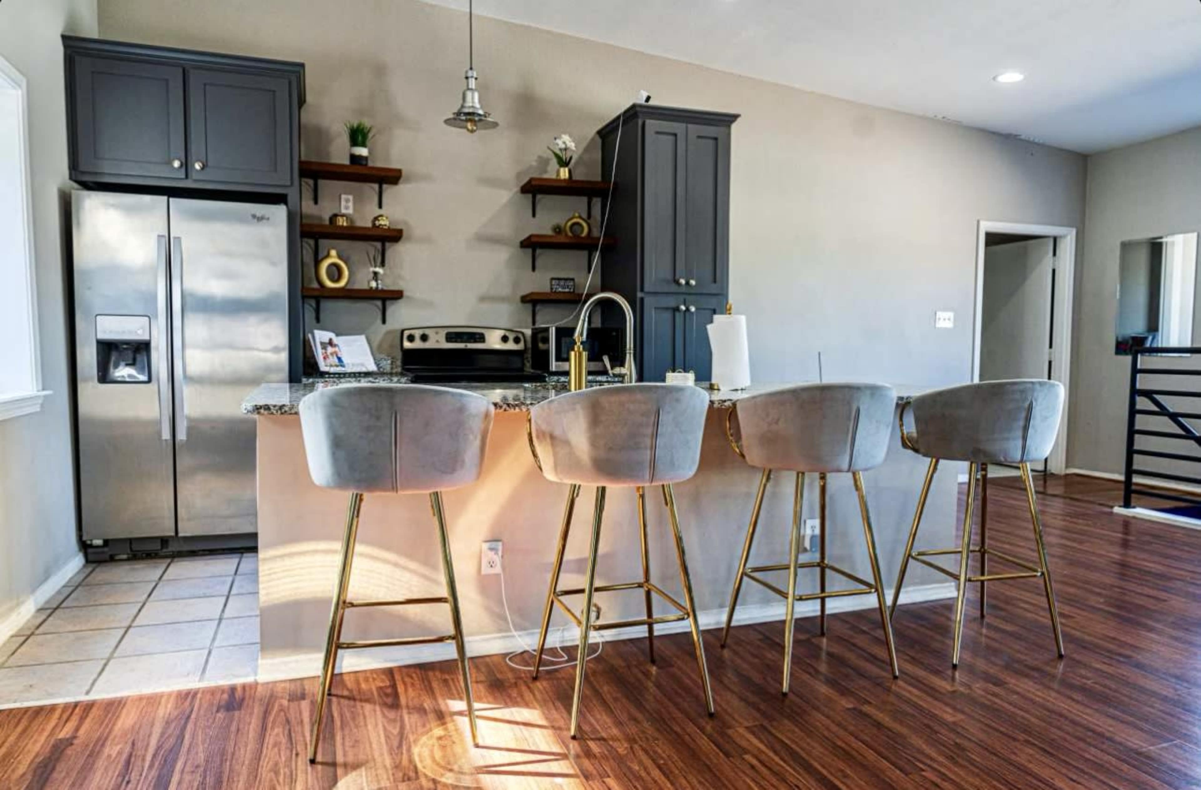 A modern kitchen features a granite island with four high-backed stools, stainless steel appliances, and dark cabinetry.