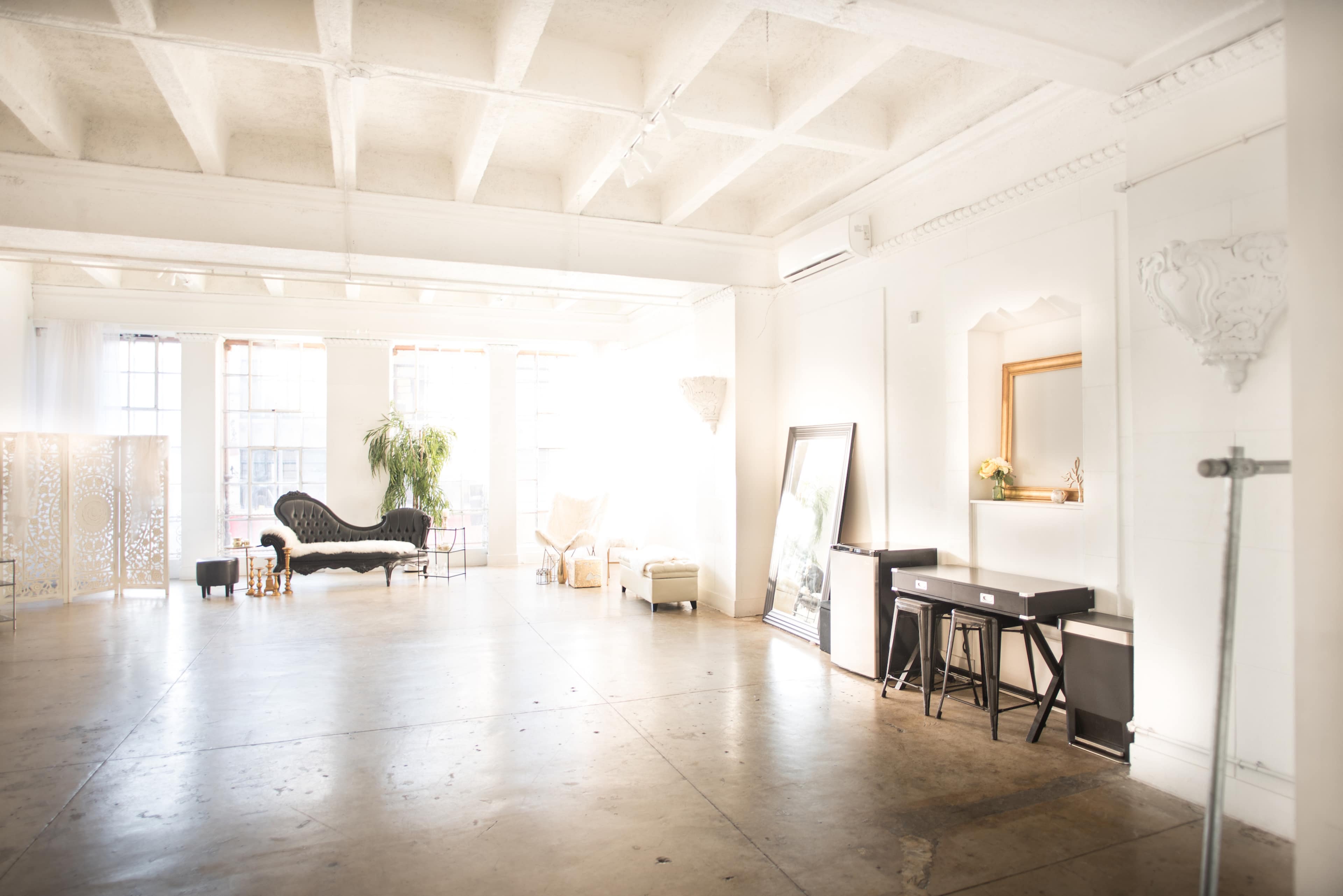 A spacious, brightly lit interior with high ceilings, featuring a mix of furniture including a black sofa, a white armchair, and a small table against the wall.