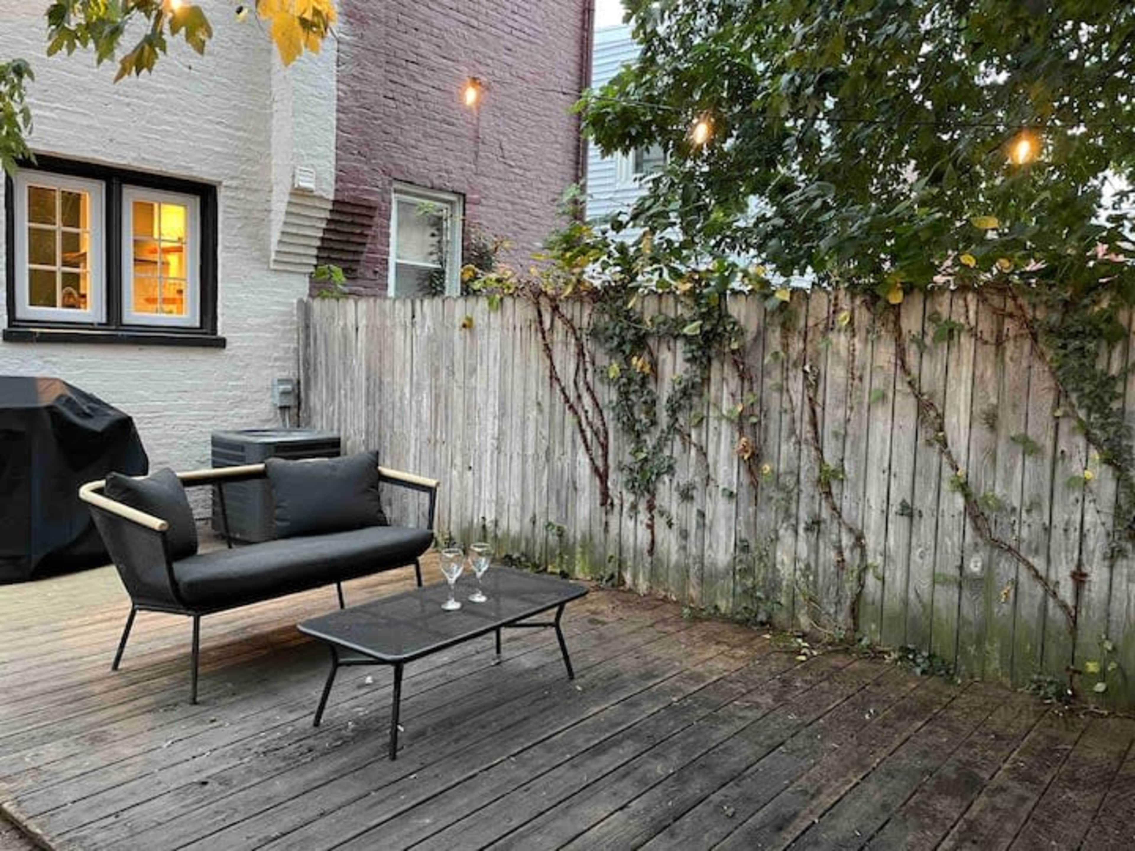 A small outdoor seating area features a black sofa and a glass table on a wooden deck, surrounded by a wooden fence with climbing vines.