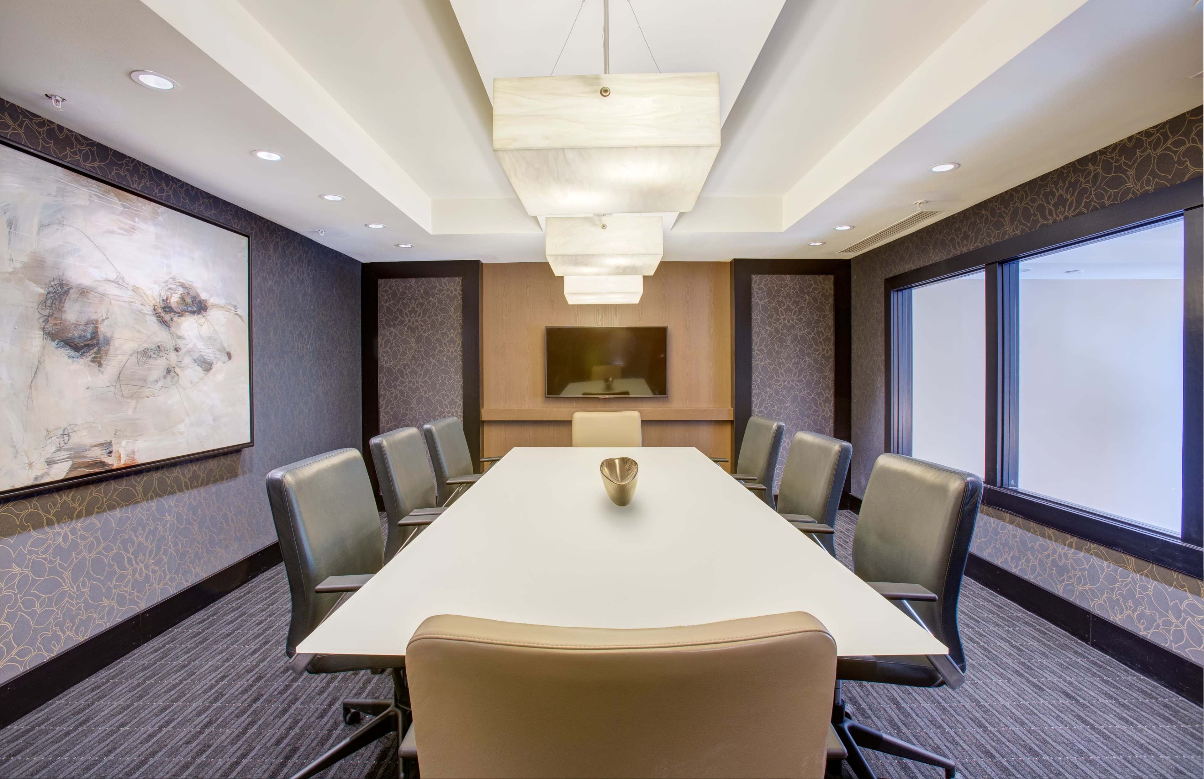 A modern conference room features a long white table surrounded by black leather chairs, with a decorative artwork on the wall and a television mounted on a wood panel.