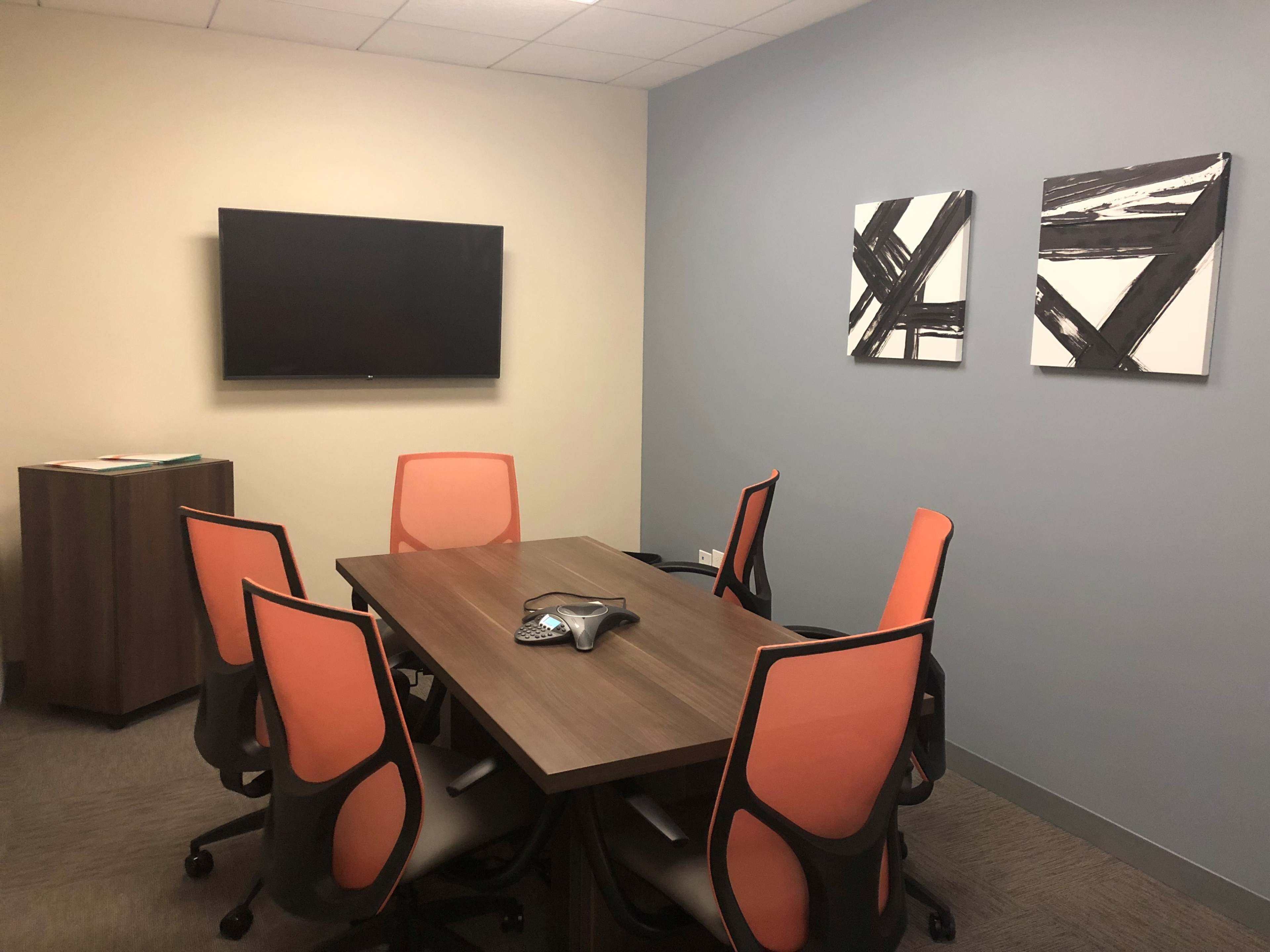 A conference room equipped with a wooden table surrounded by six orange and black chairs, a wall-mounted television, and two abstract framed artworks.