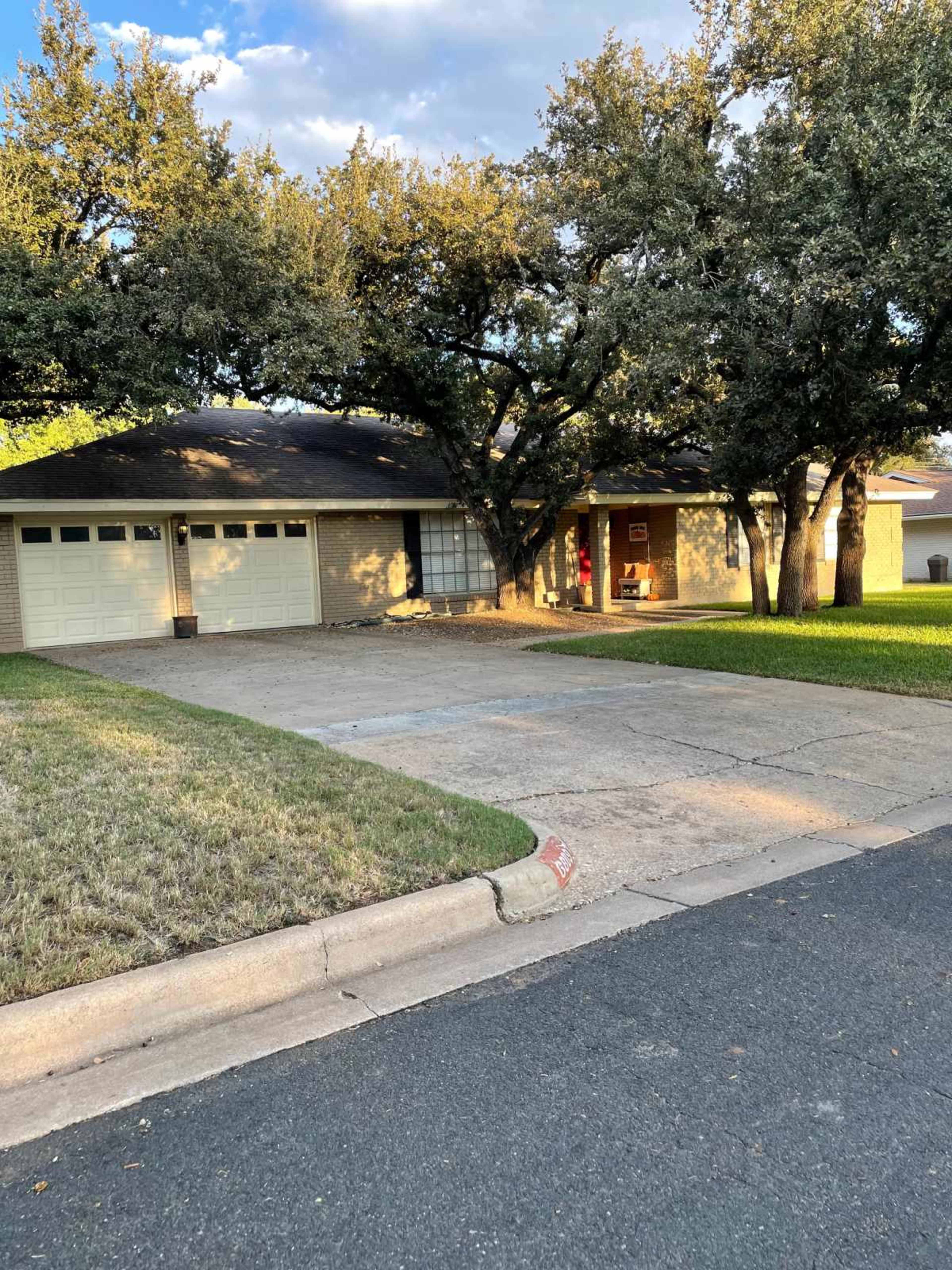 A single-story house with a two-car garage is set back from the street, surrounded by large trees and a grassy yard.