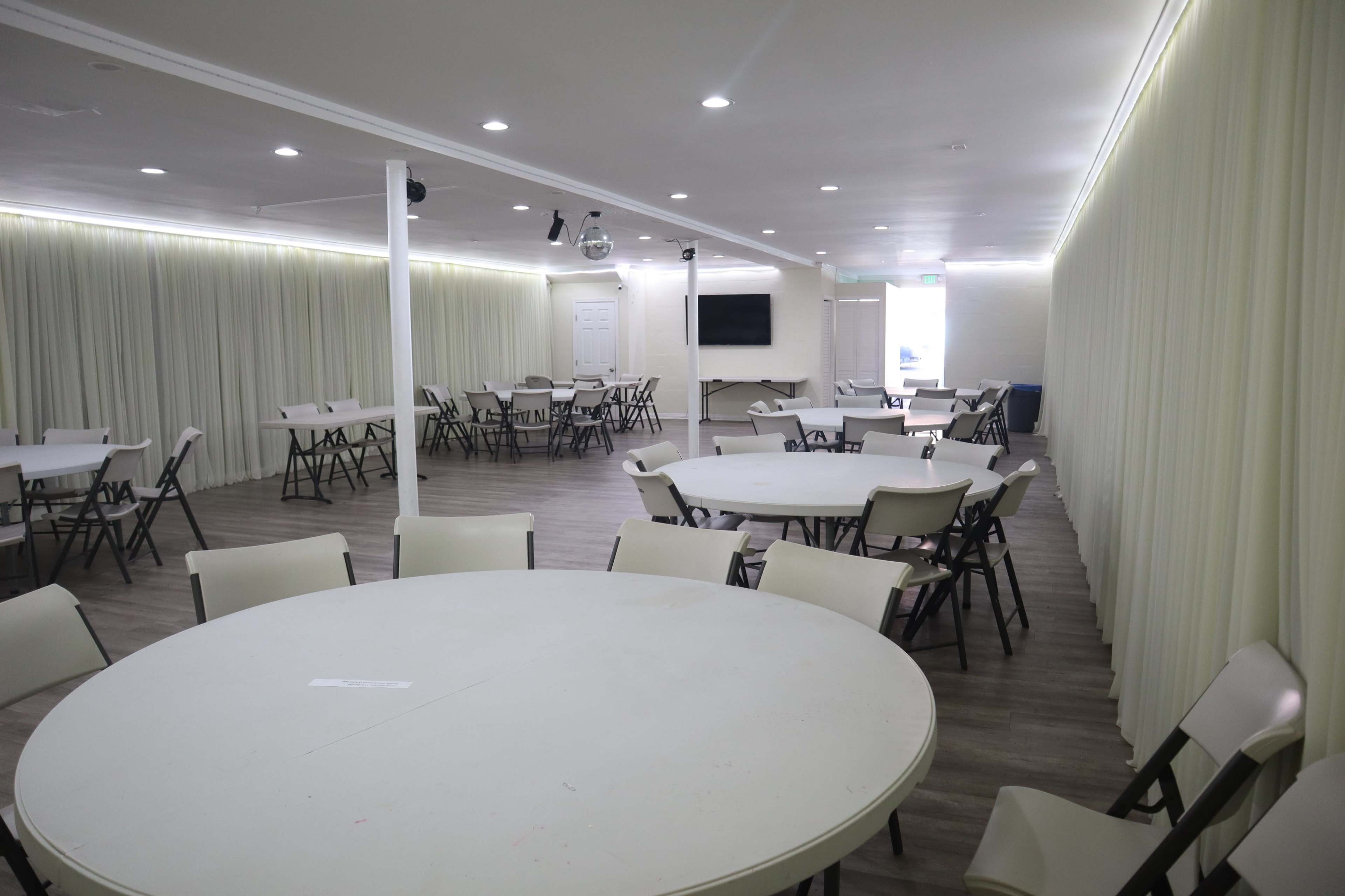 An empty event room with round tables and chairs arranged throughout, illuminated by overhead lights and characterized by light-colored curtains along the walls.