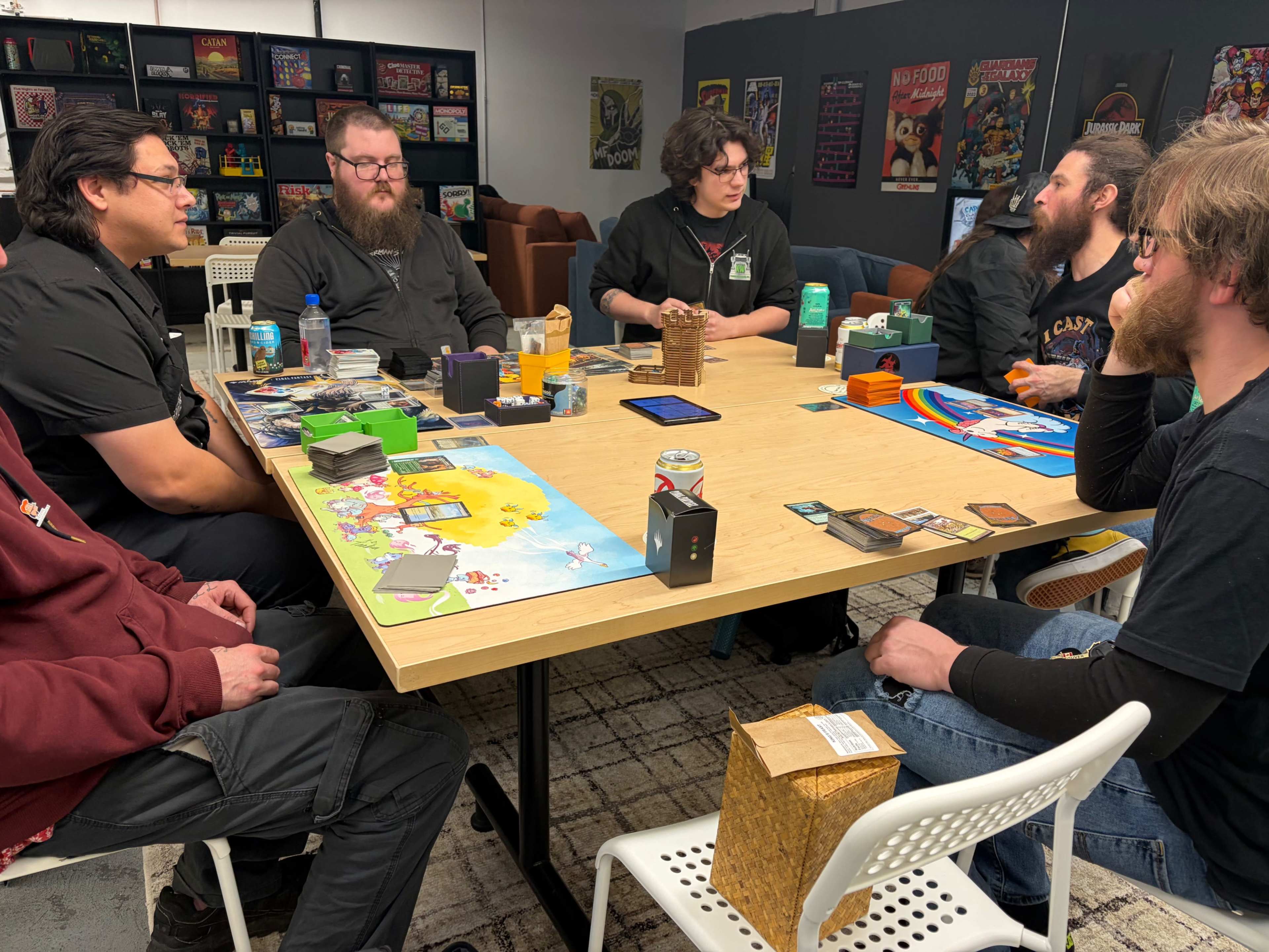 A group of seven people is sitting around a table playing tabletop games, with various game boxes and cards spread out in front of them.