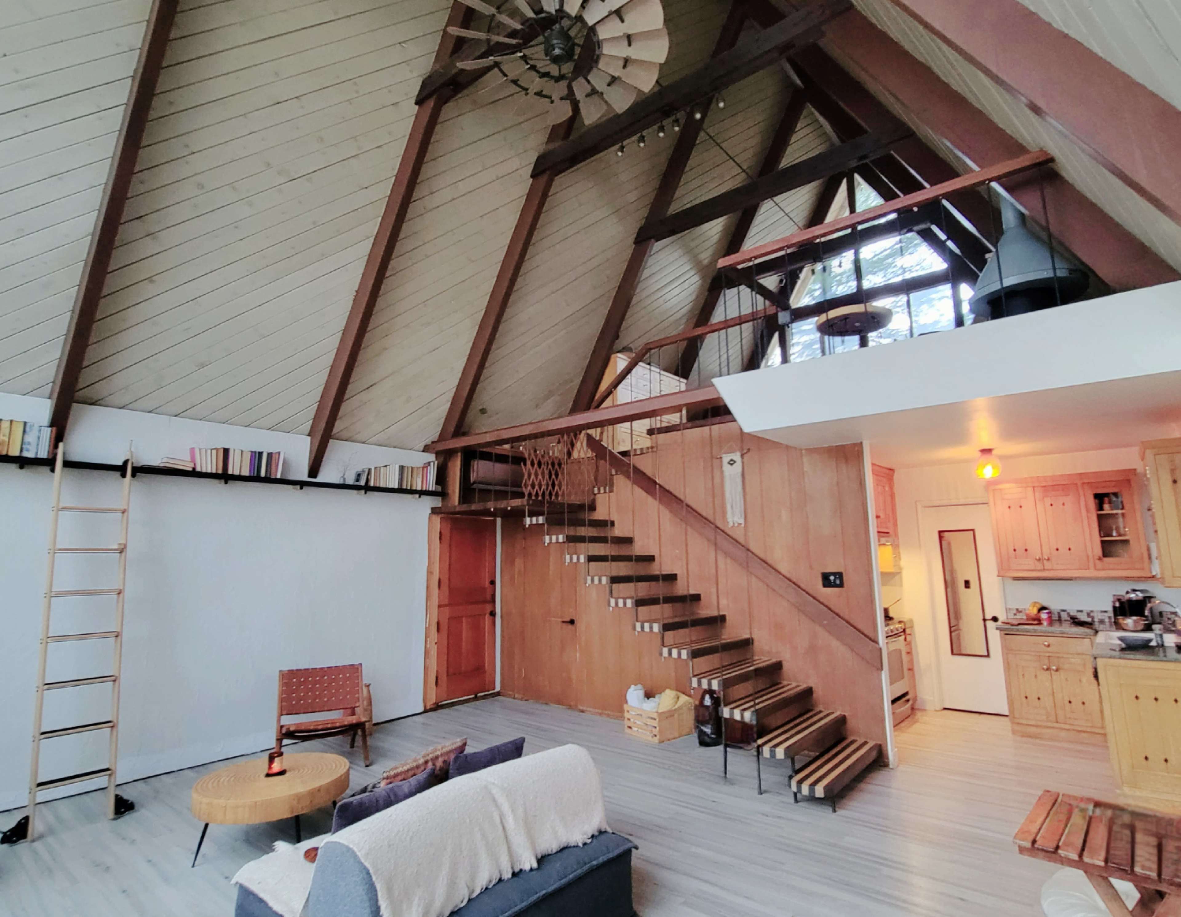 A spacious interior of an A-frame house with a wooden staircase, a seating area, and a kitchen visible in the background.