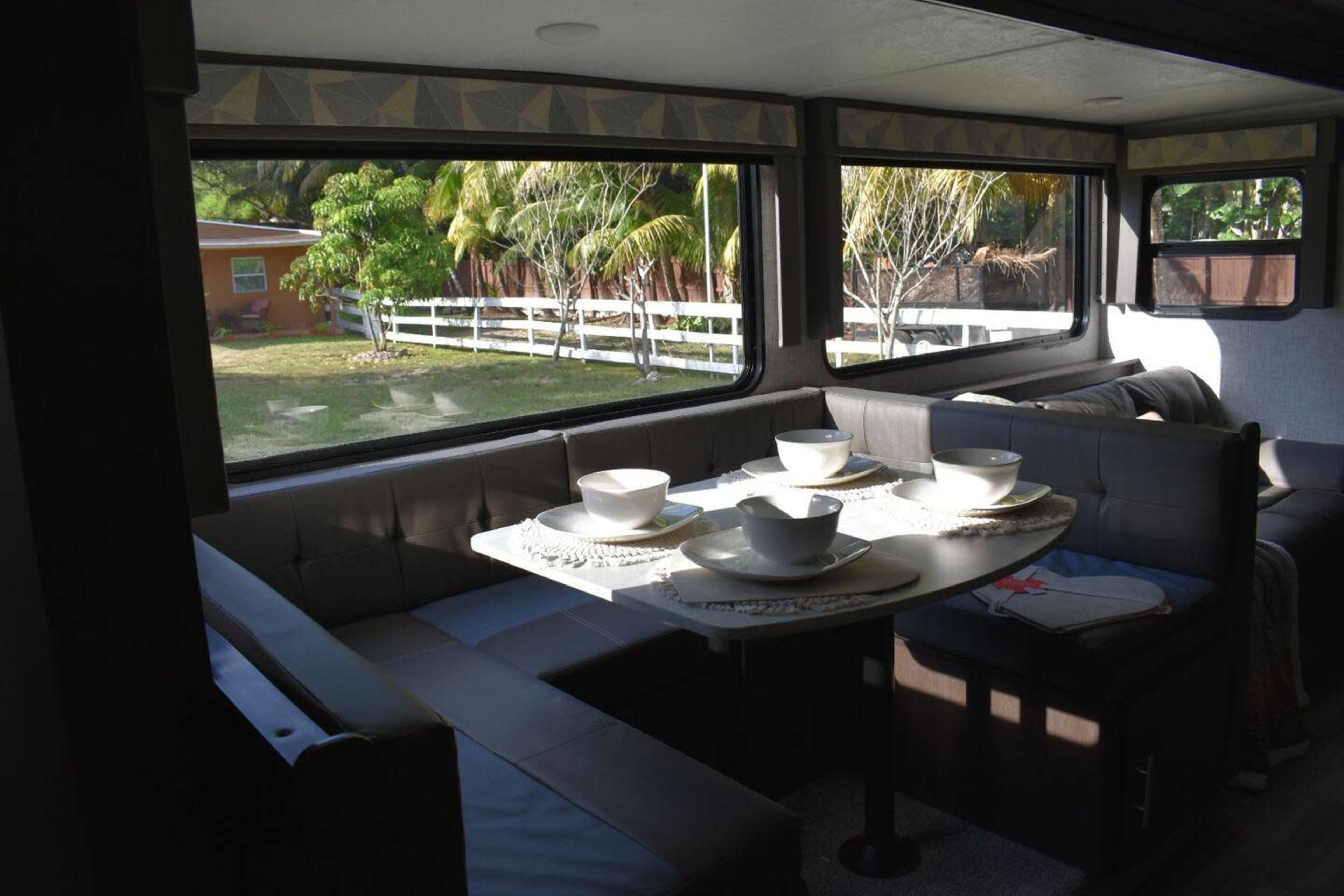 The image shows a dining area inside a modern camper with a table set for four, featuring bowls and placemats, and large windows providing a view of greenery outside.