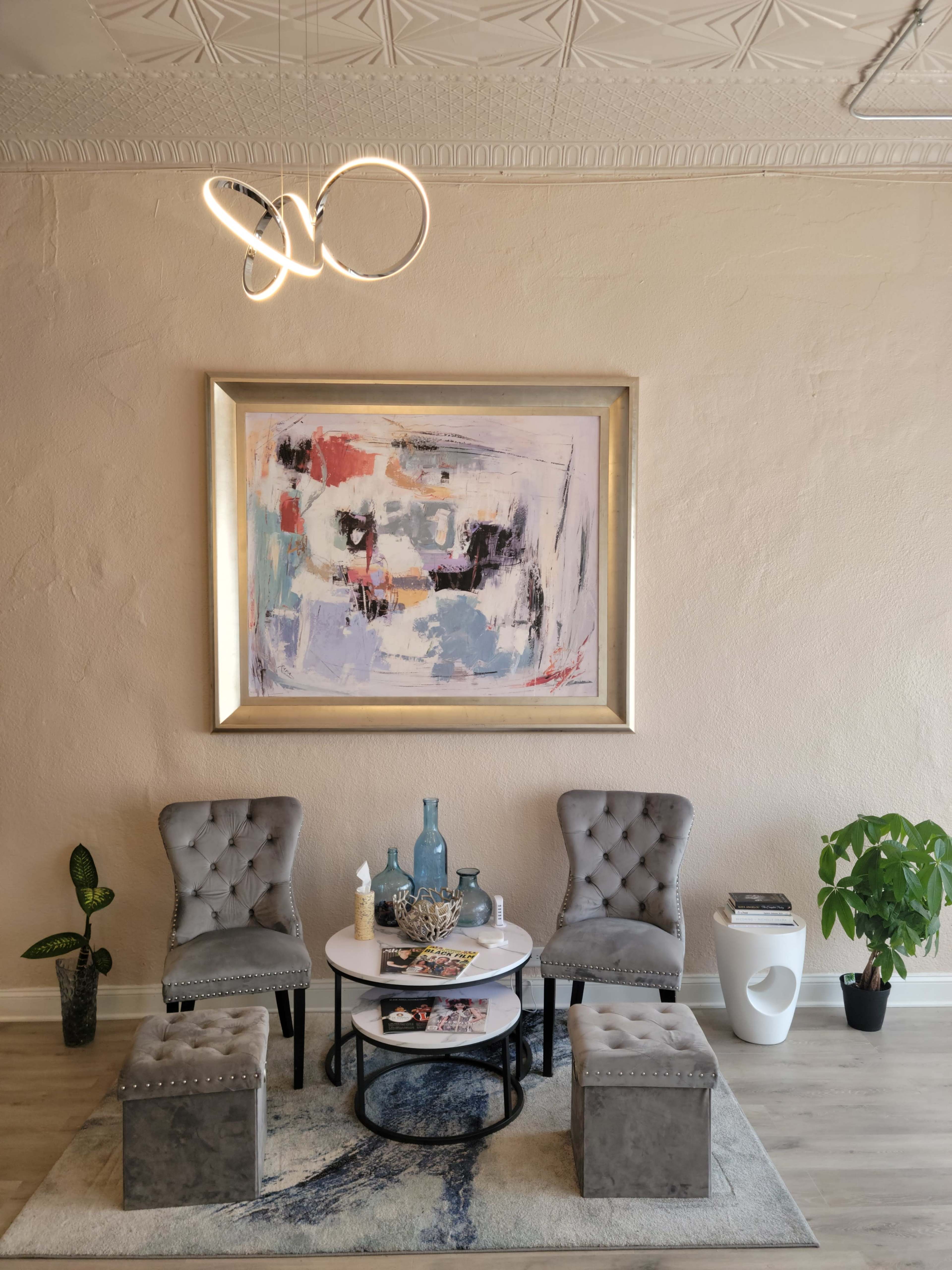 A modern lounge area features two tufted gray chairs and a round coffee table, accented by a colorful abstract painting on the wall and decorative plants.