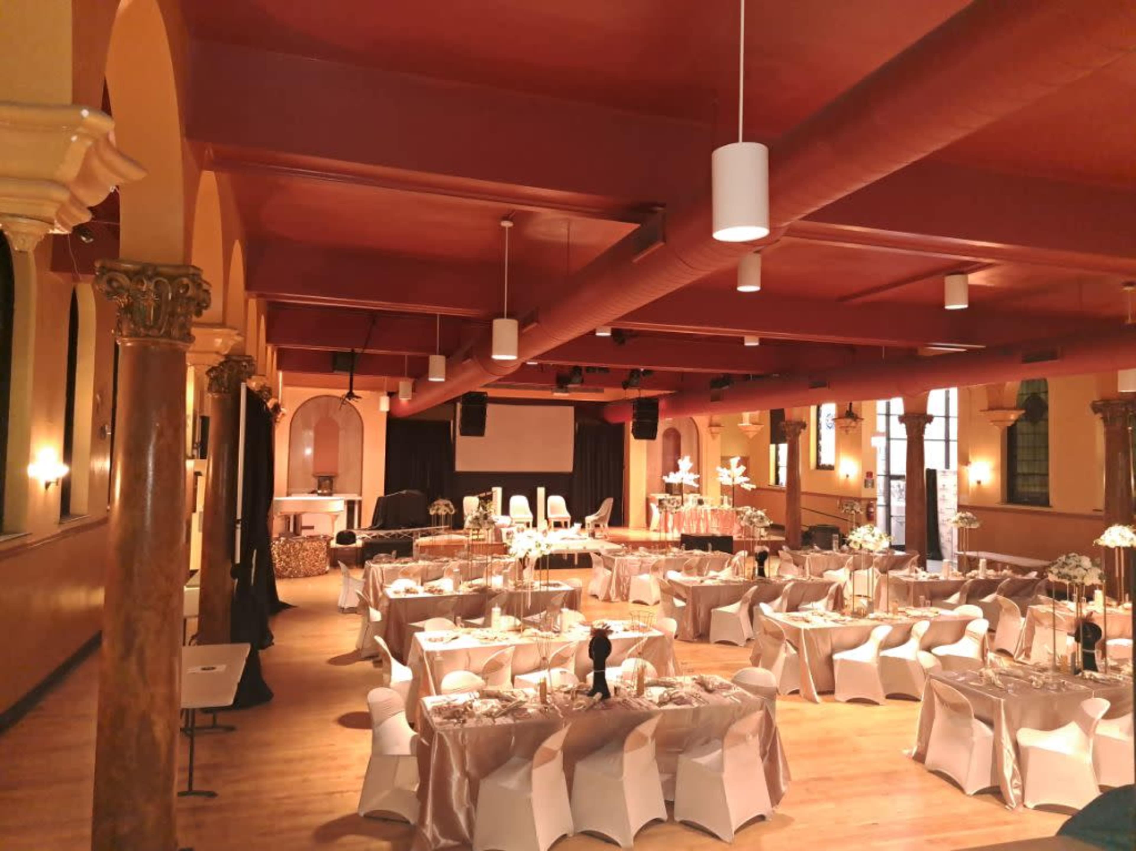 A spacious banquet hall arranged for an event, featuring elegantly set tables with white tablecloths and chairs, under warm lighting and decorative columns.