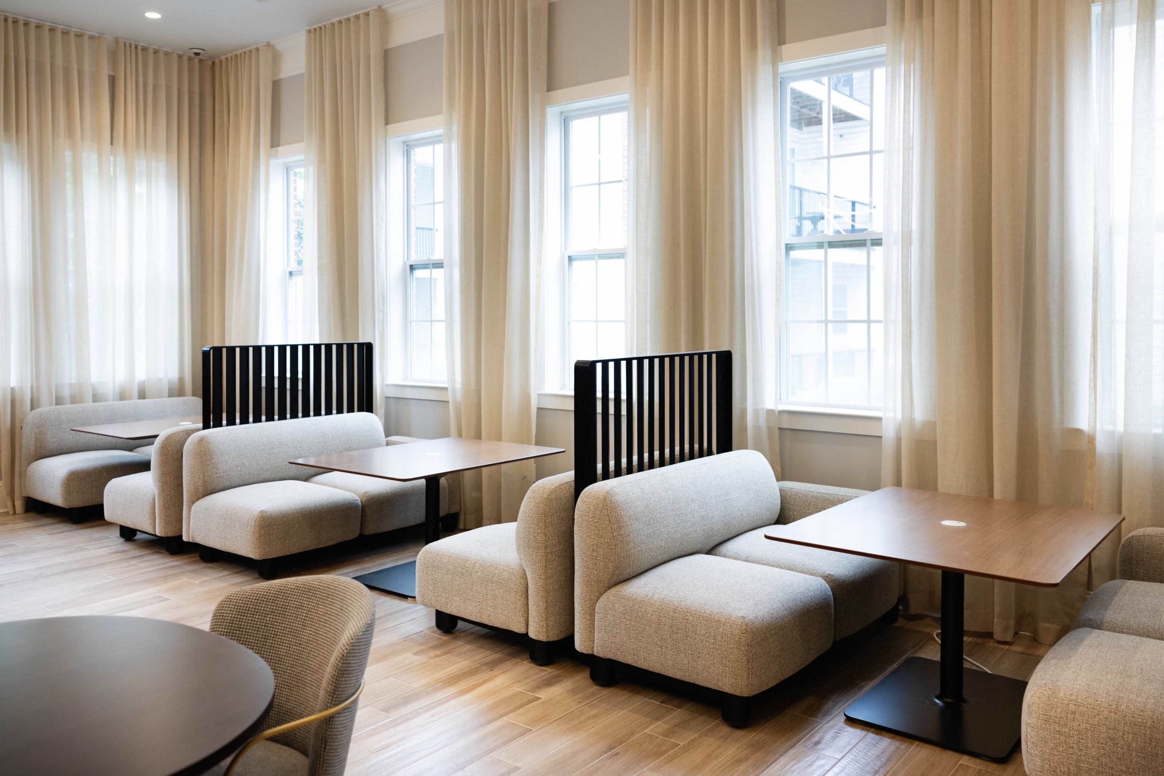The image shows a contemporary seating area featuring light-colored, modular couches and wooden tables arranged near large windows with sheer curtains.