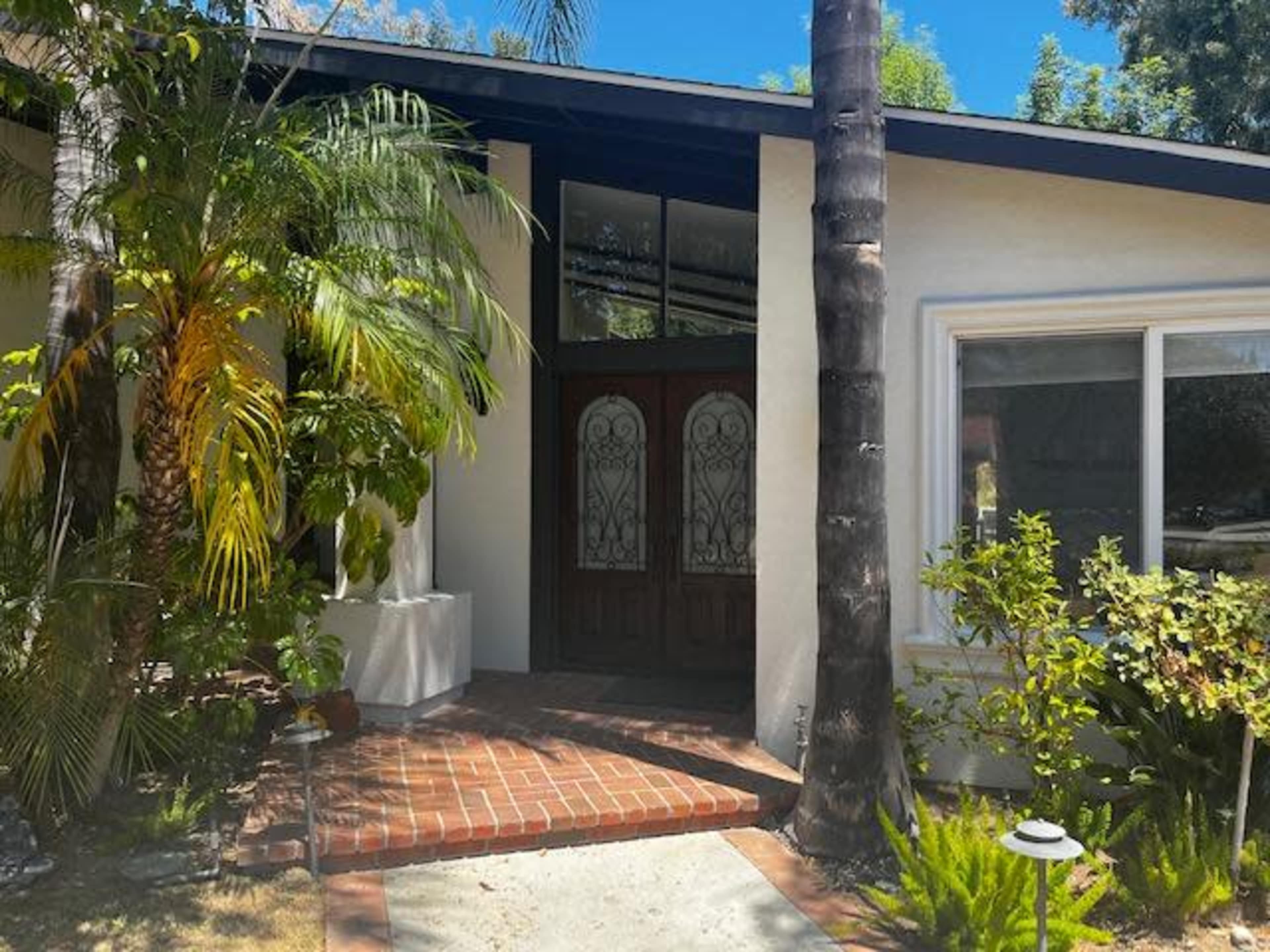 A house entrance with double wooden doors, flanked by palm trees and surrounded by landscaped greenery.
