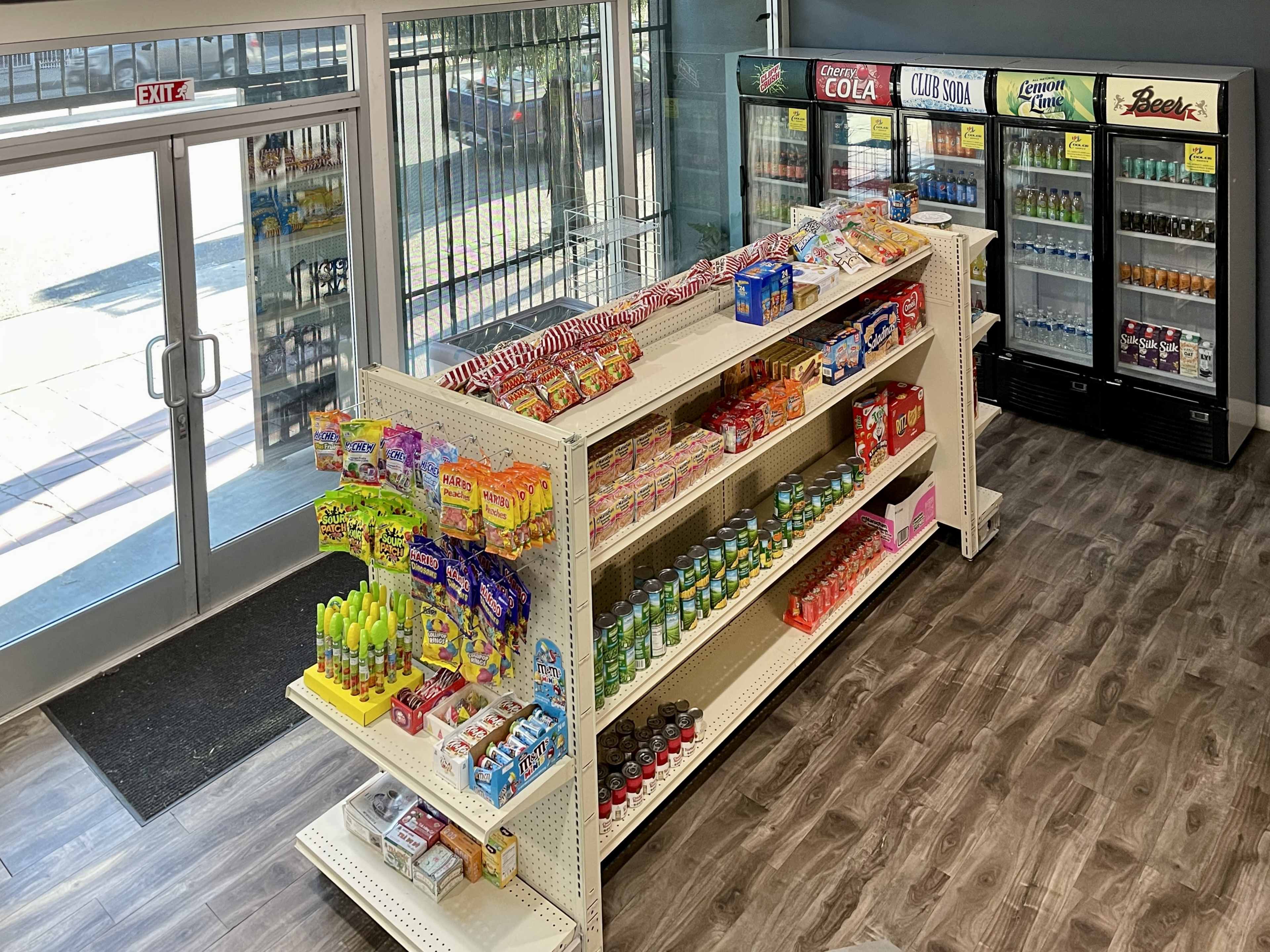 A store interior with an aisle of shelves stocked with various snacks and beverages, facing large glass doors that provide an exit.