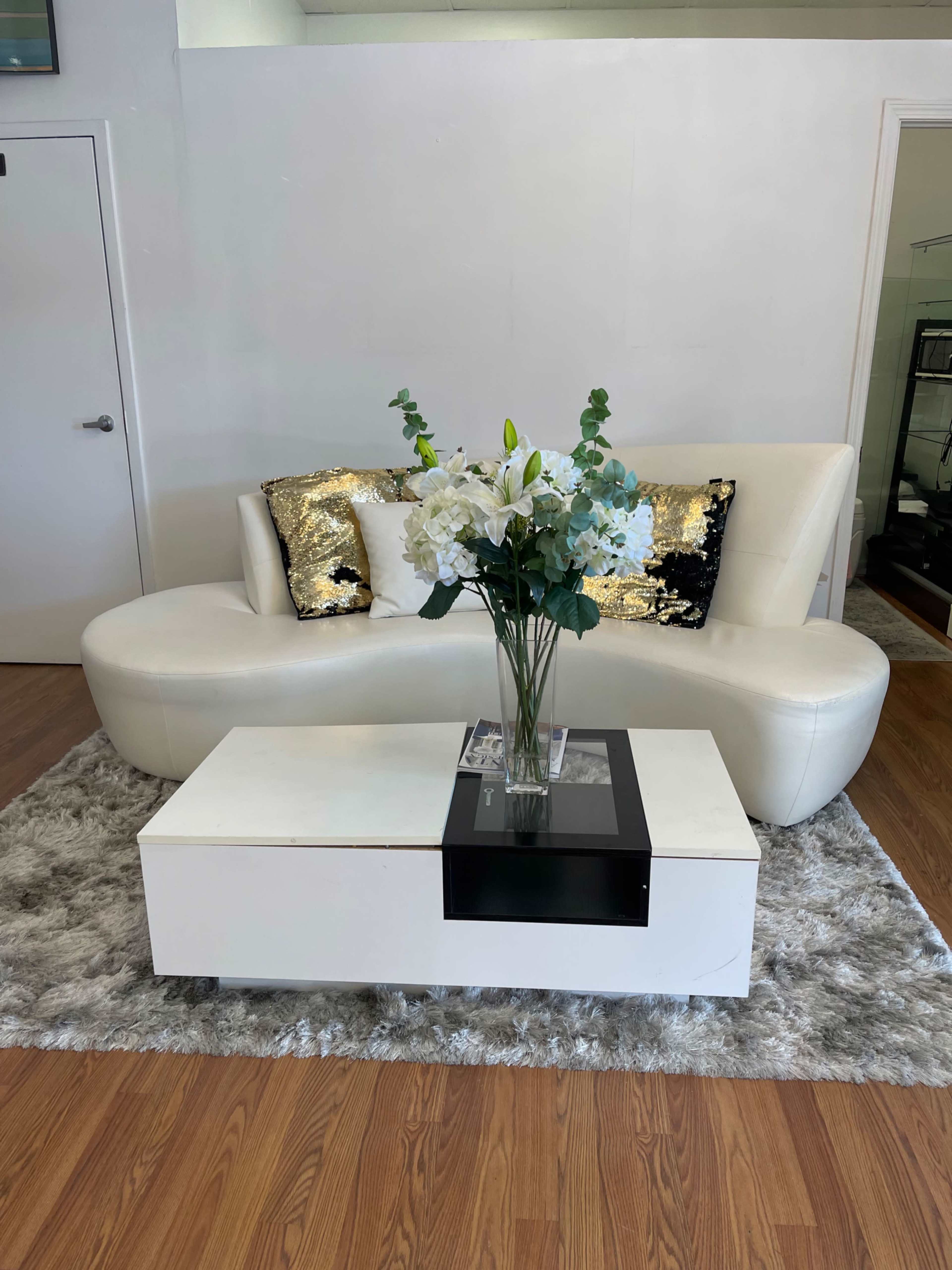 A white sofa with decorative pillows is positioned next to a modern coffee table adorned with a vase of flowers, set on a plush rug in a minimalist space.