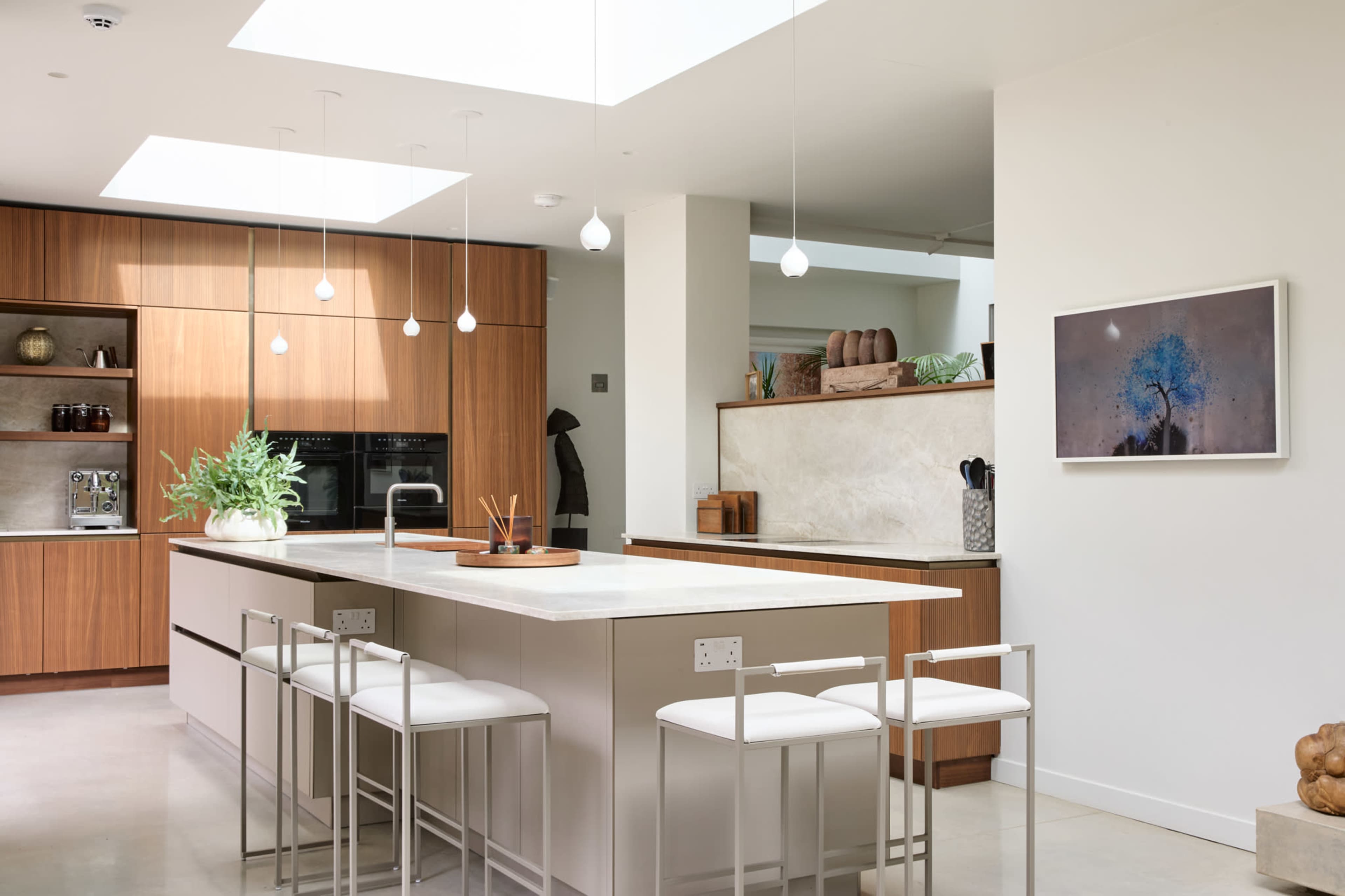 The image shows a modern kitchen with wooden cabinetry, a central island with seating, and large overhead skylights providing natural light.