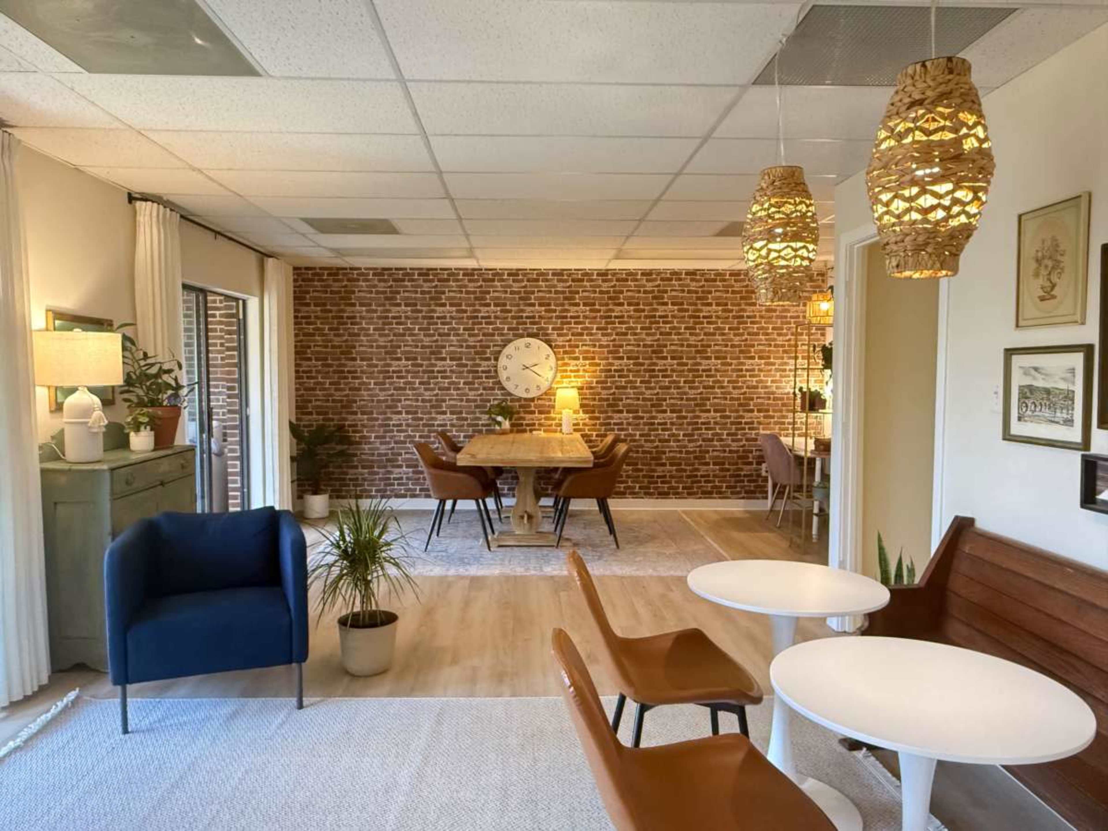 The image shows a modern interior space with a dining area featuring a wooden table, chairs, and decorative lighting, along with a small seating area that includes a couch and round tables.