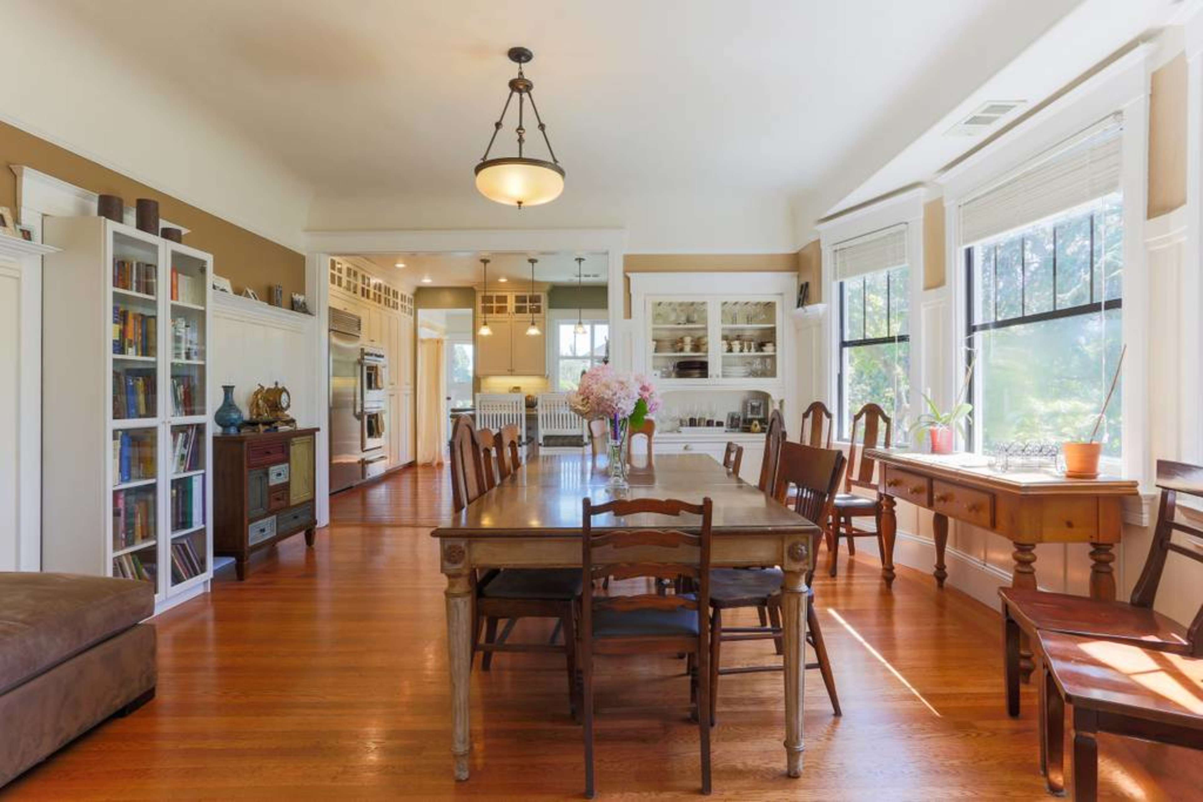 A spacious dining area with a long wooden table surrounded by chairs, illuminated by natural light from large windows, and a view into a kitchen area in the background.