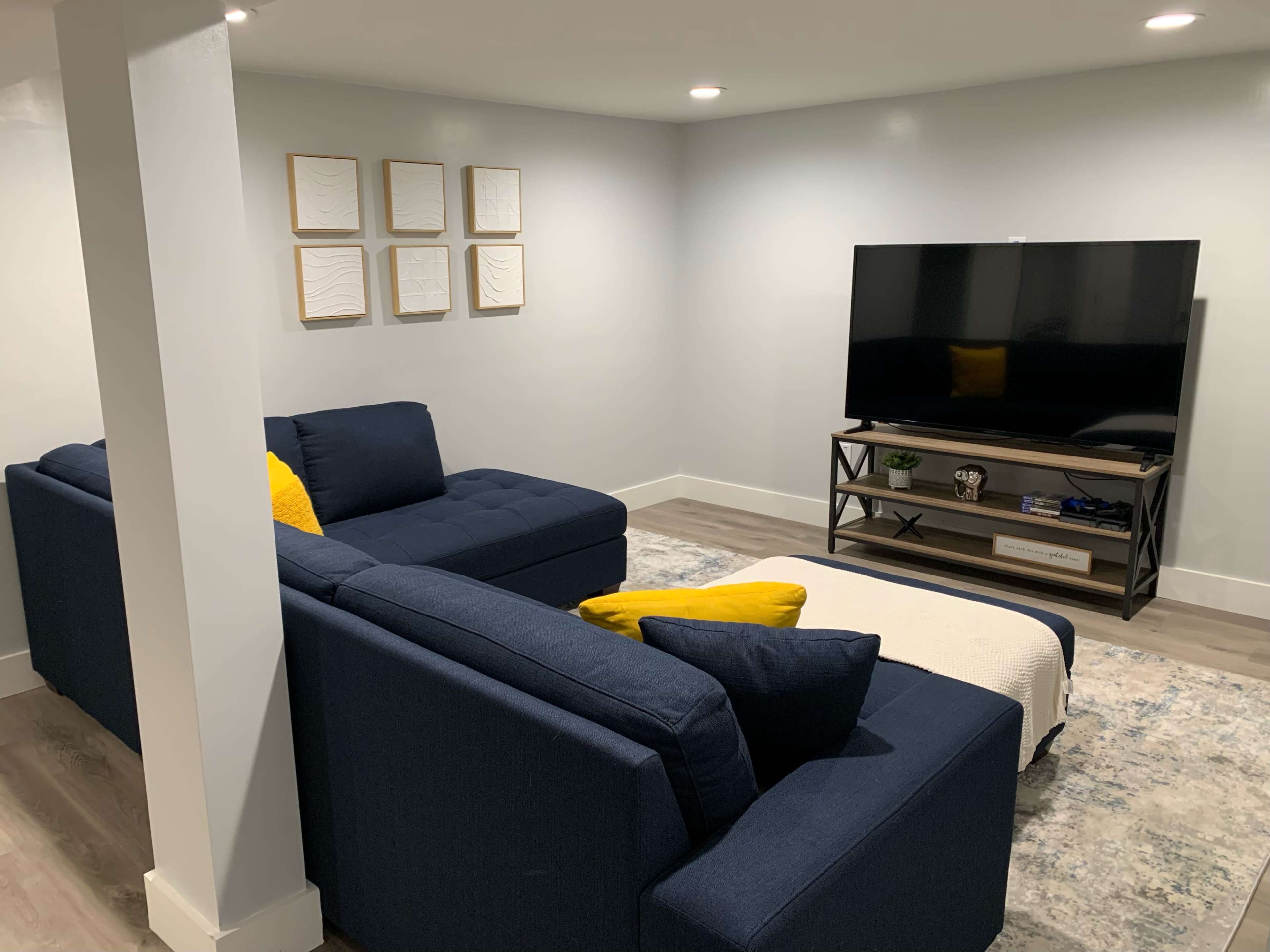 A modern basement living room features a dark blue sectional sofa, a wooden TV stand with a television, and framed artworks on the wall.
