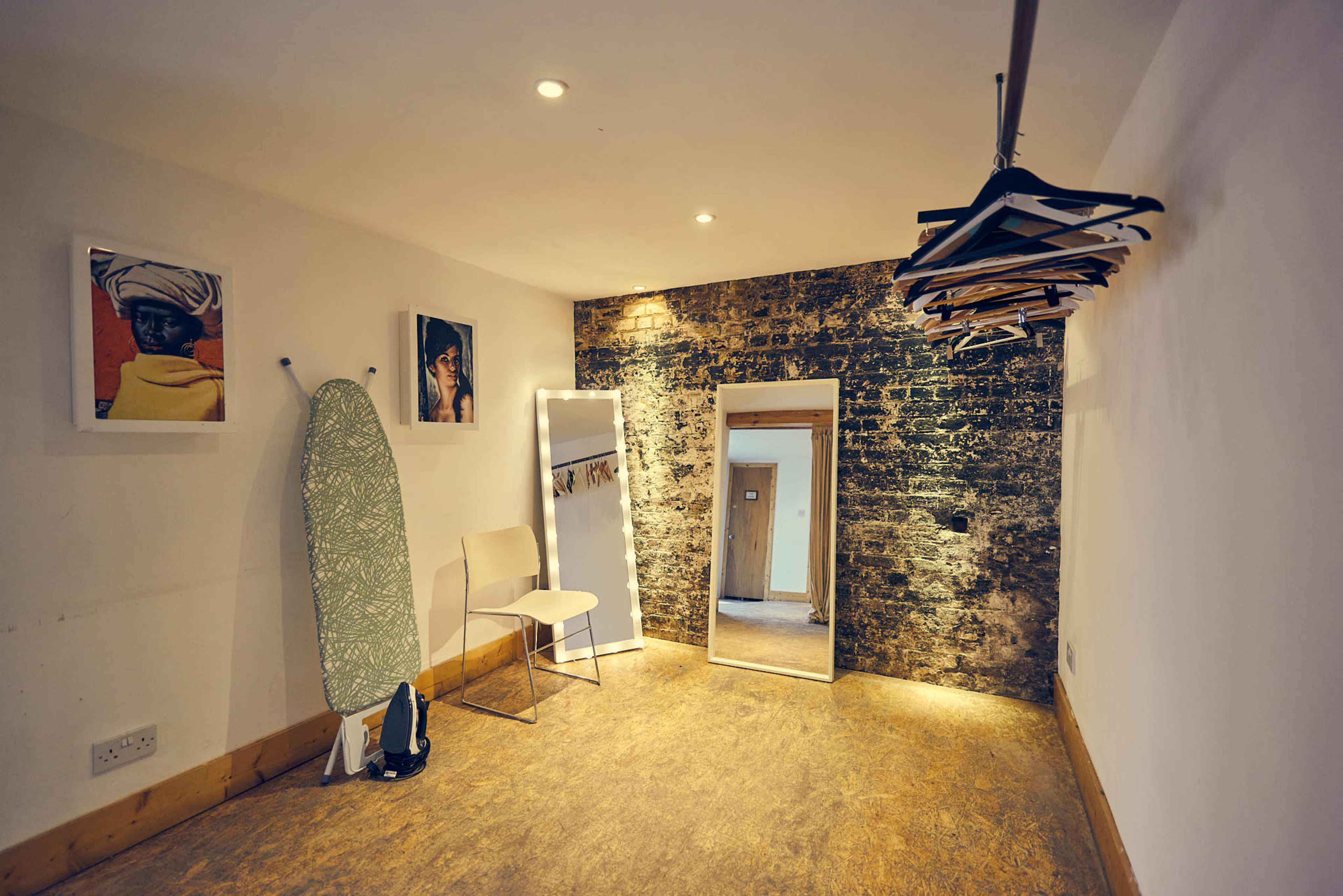 A minimalist room featuring a chair, an ironing board, a full-length mirror, a clothing rack with hangers, and two framed photographs on the wall.