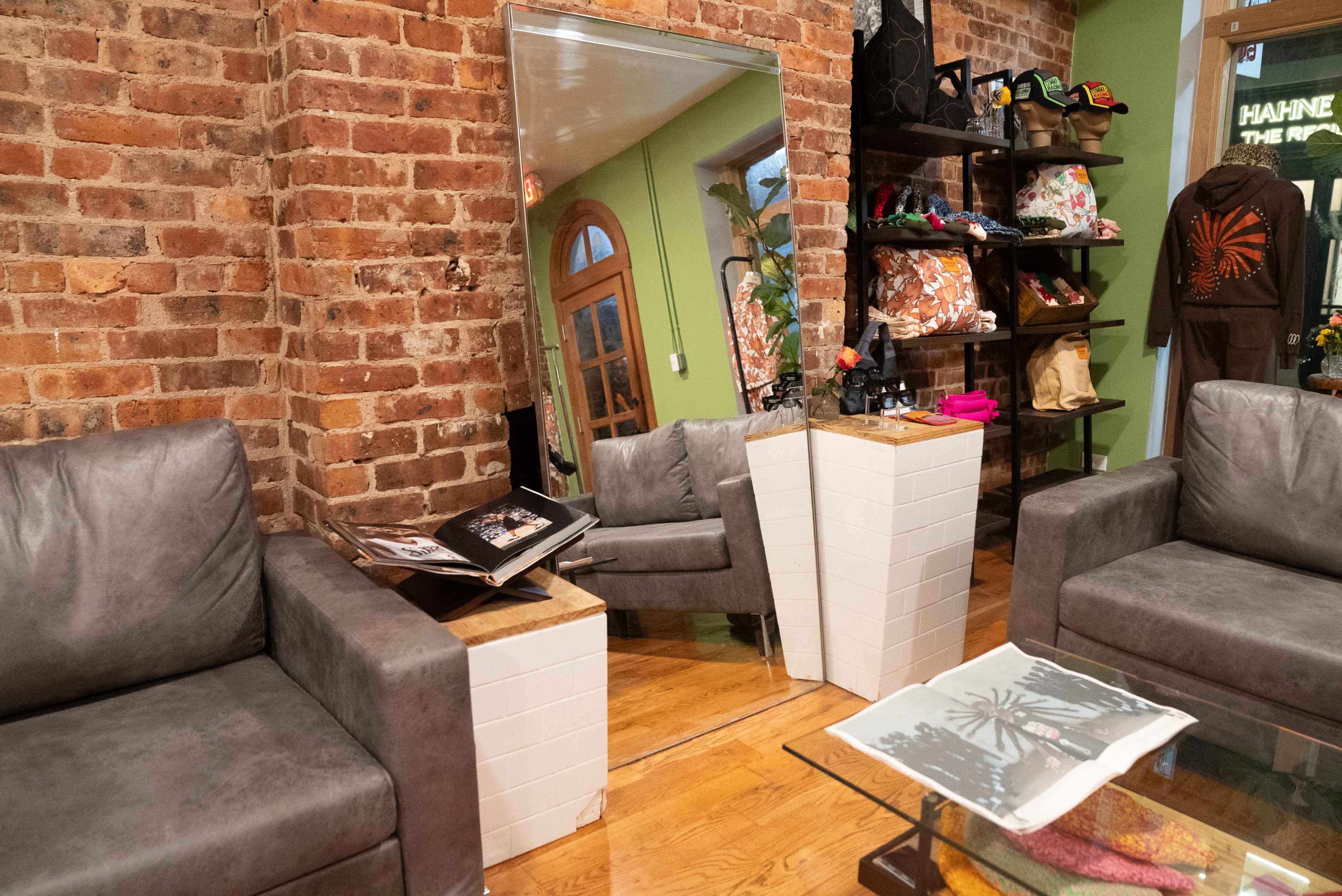 The image shows a cozy boutique with exposed brick walls, two gray sofas, a glass coffee table, and shelves displaying various accessories.