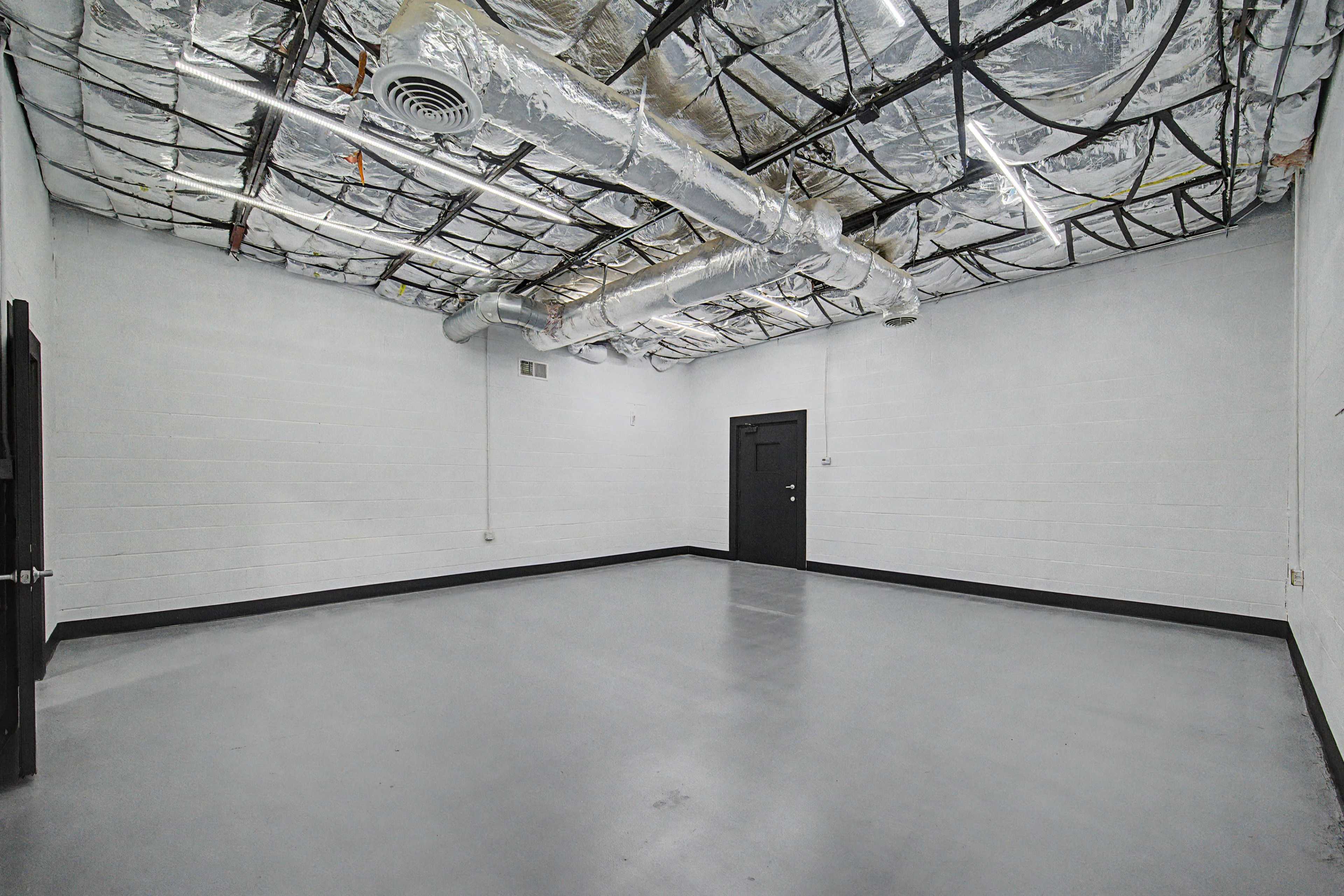 The image shows an empty room with gray walls, a smooth gray floor, and exposed metallic ductwork on the ceiling.