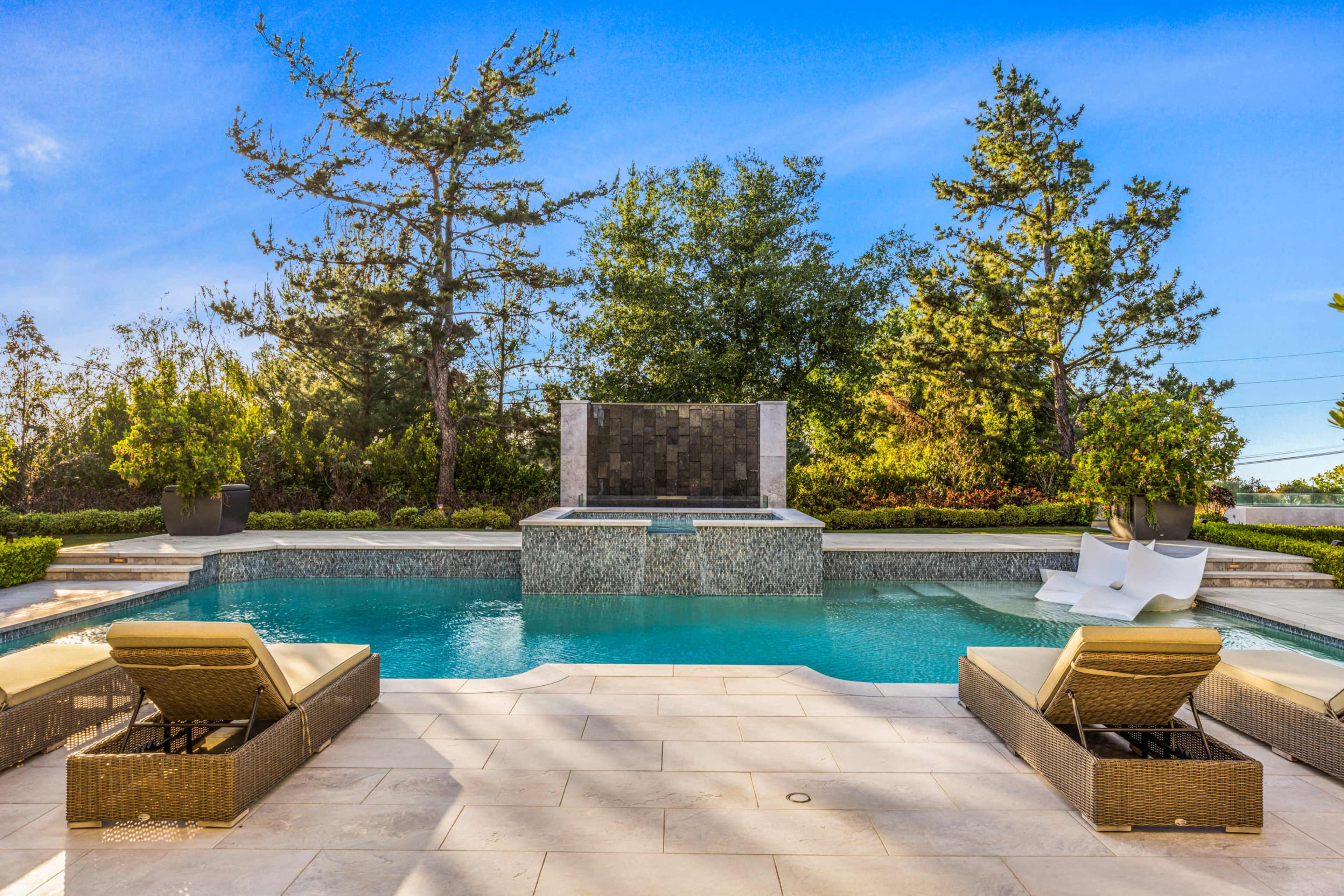 A modern pool area features two lounge chairs beside a tranquil water feature and surrounding greenery.