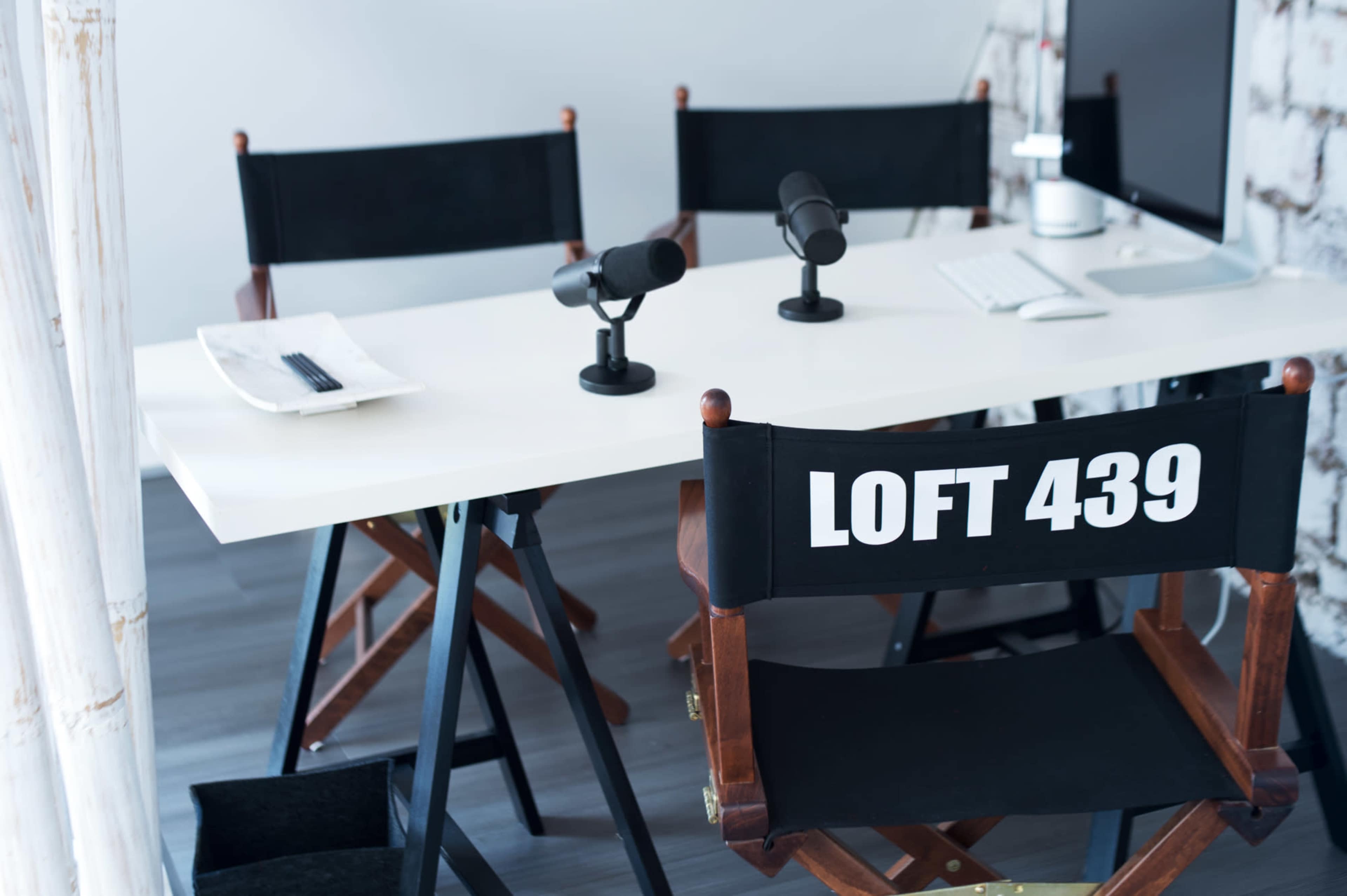 A modern workspace features a white table with two microphones and a computer, complemented by director's chairs labeled "LOFT 439."