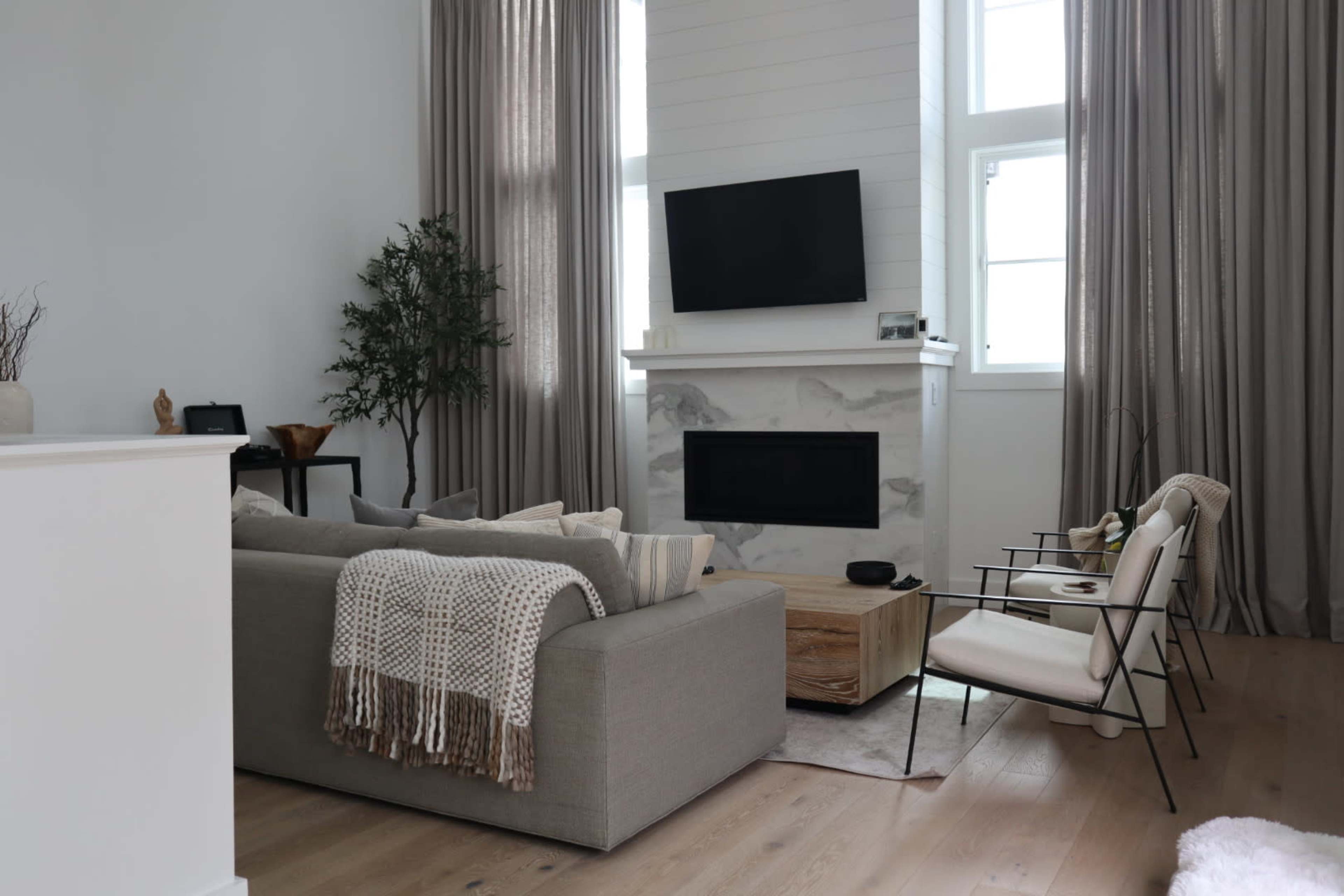 A modern living room featuring a gray sofa, a wooden coffee table, a fireplace with a white surround, and large windows covered with long, light-colored curtains.