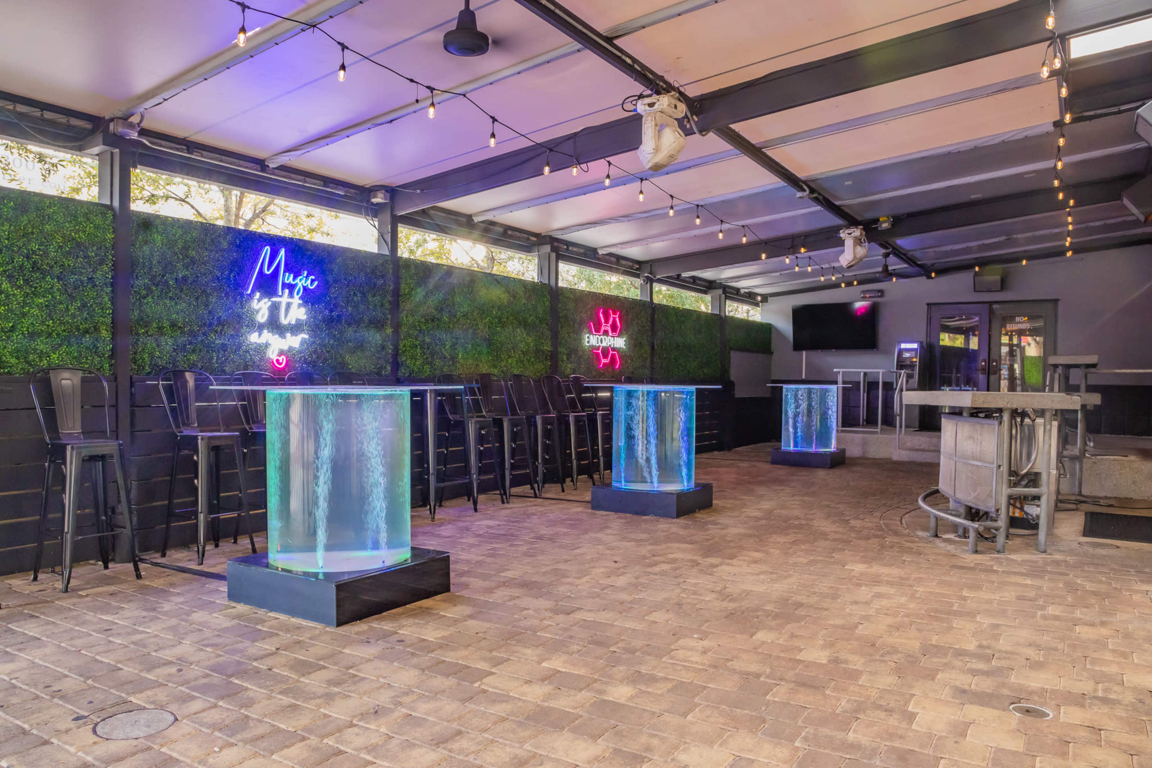 The image shows a modern outdoor bar area featuring illuminated glass tables, bar stools, and neon signs against a backdrop of greenery.