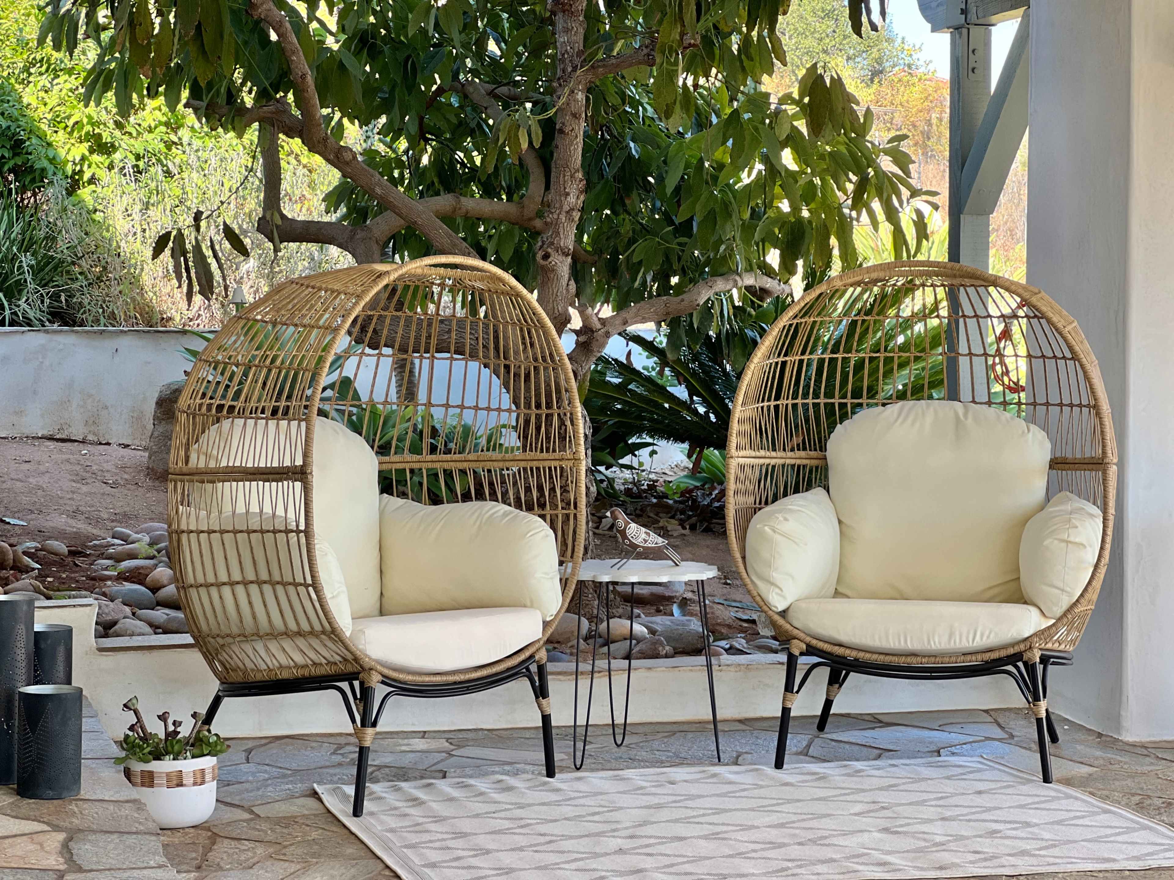 The scene features two circular rattan chairs with cushions placed next to a small side table, surrounded by greenery and stone landscaping.
