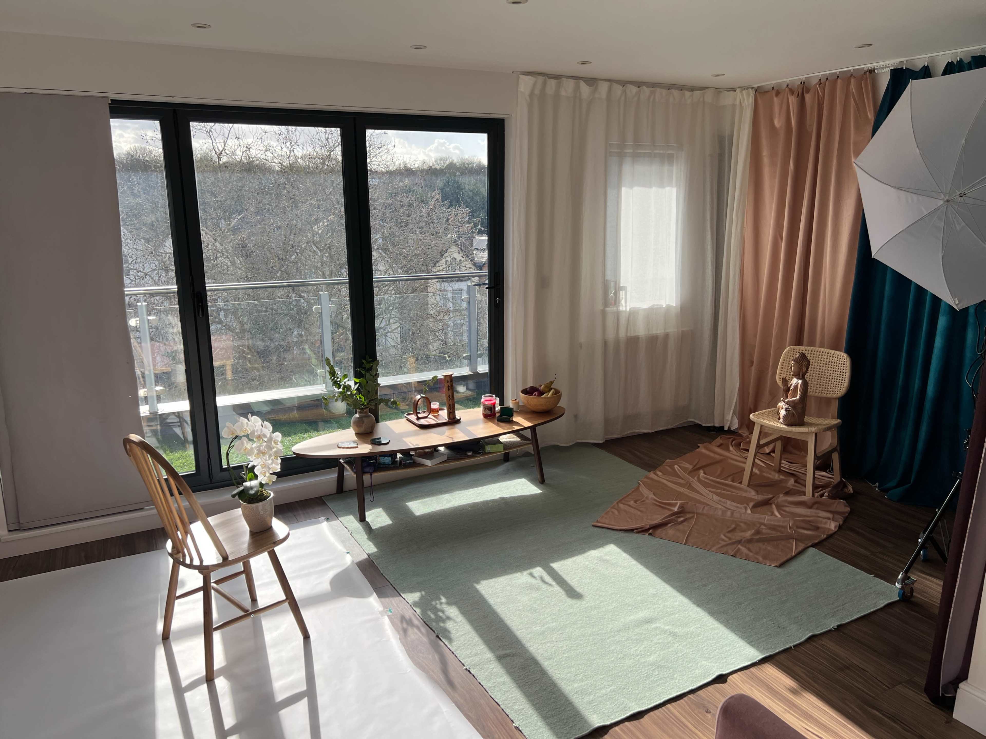 The image shows a bright interior room with large windows, a green area rug, a wooden table set with decorative items, and a small chair next to a soft brown fabric.