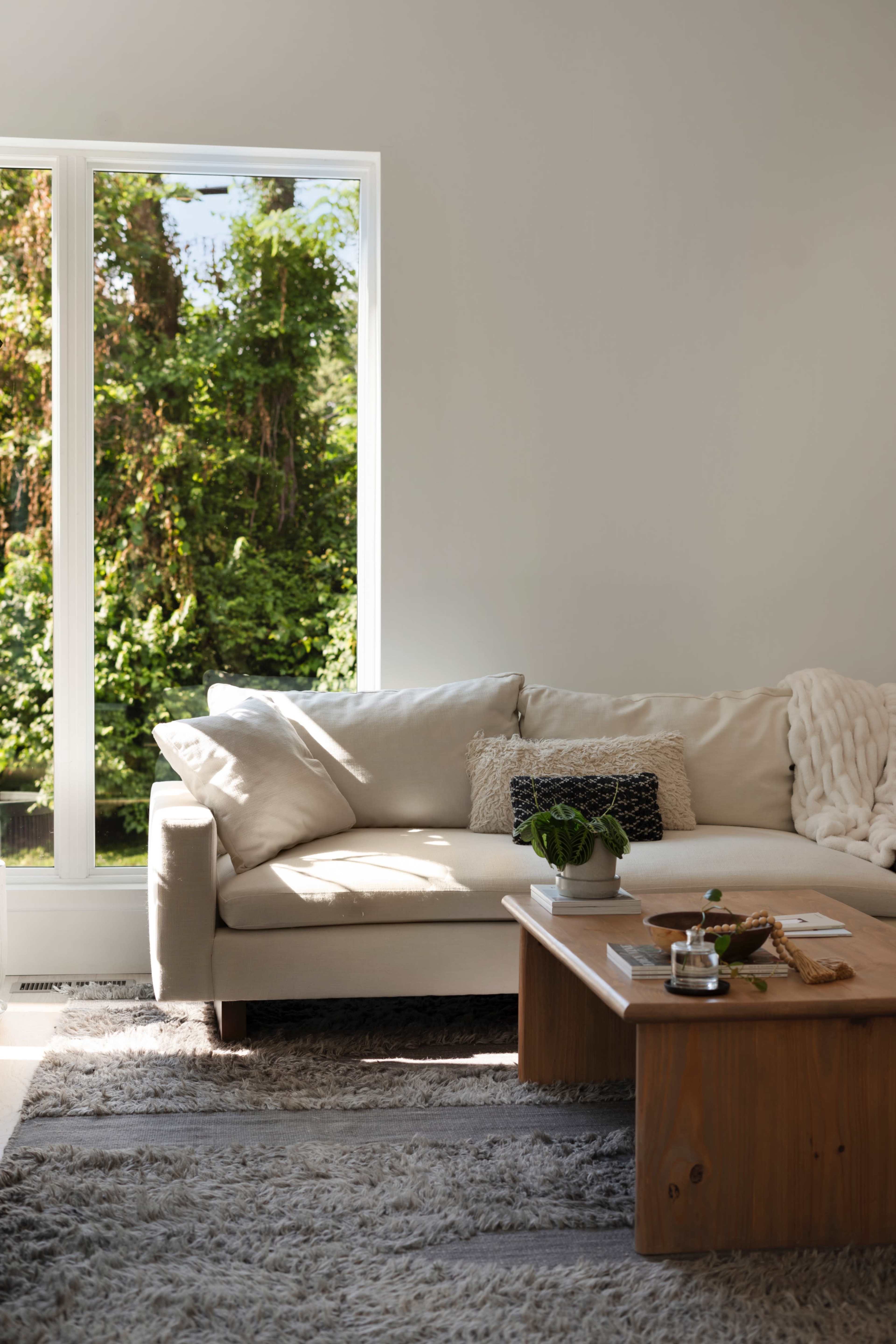 A modern living room features a light-colored sectional sofa, a wooden coffee table, and a large window showcasing lush greenery outside.