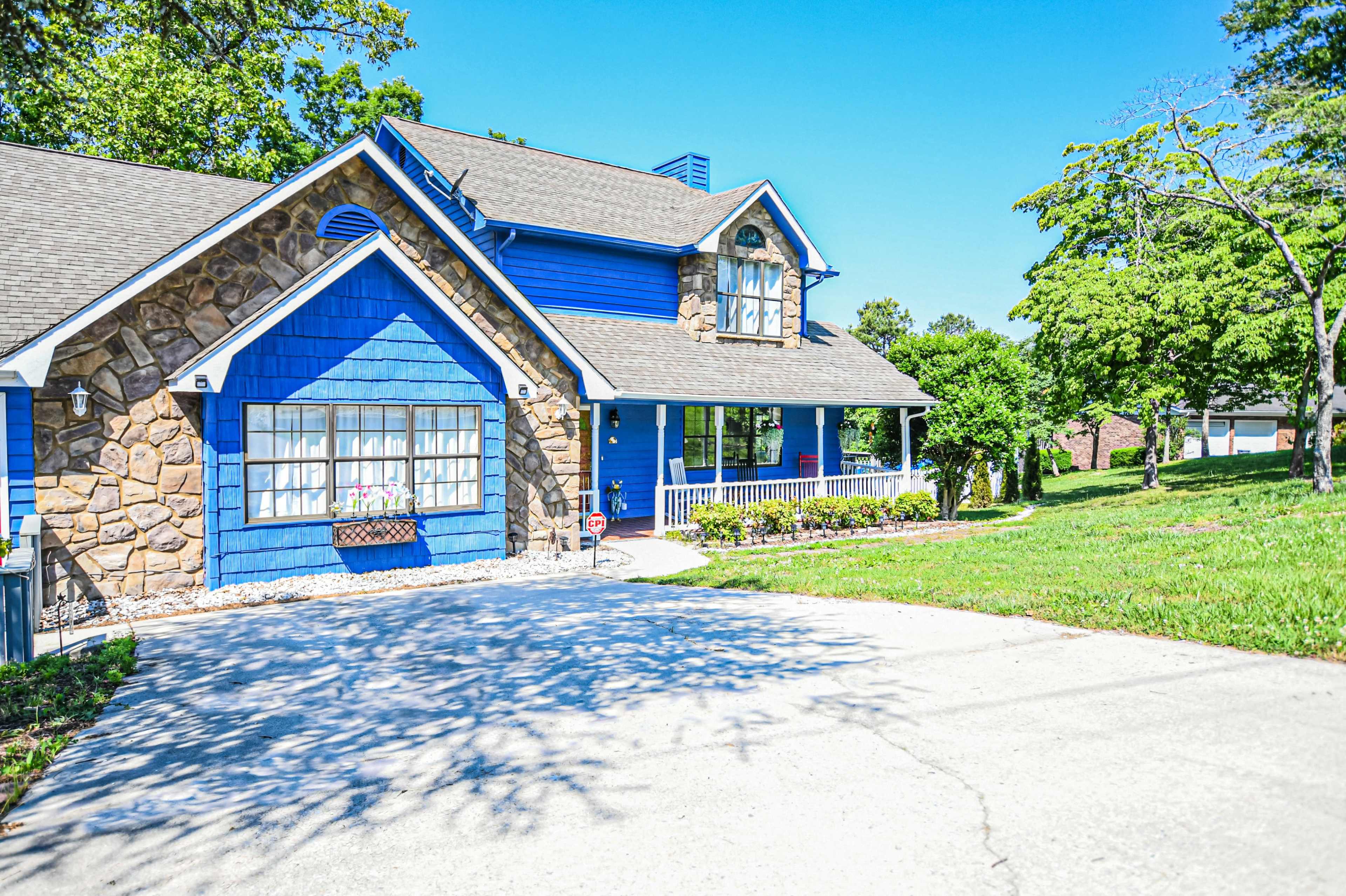 A blue and stone house with a front porch is surrounded by trees and an open lawn.
