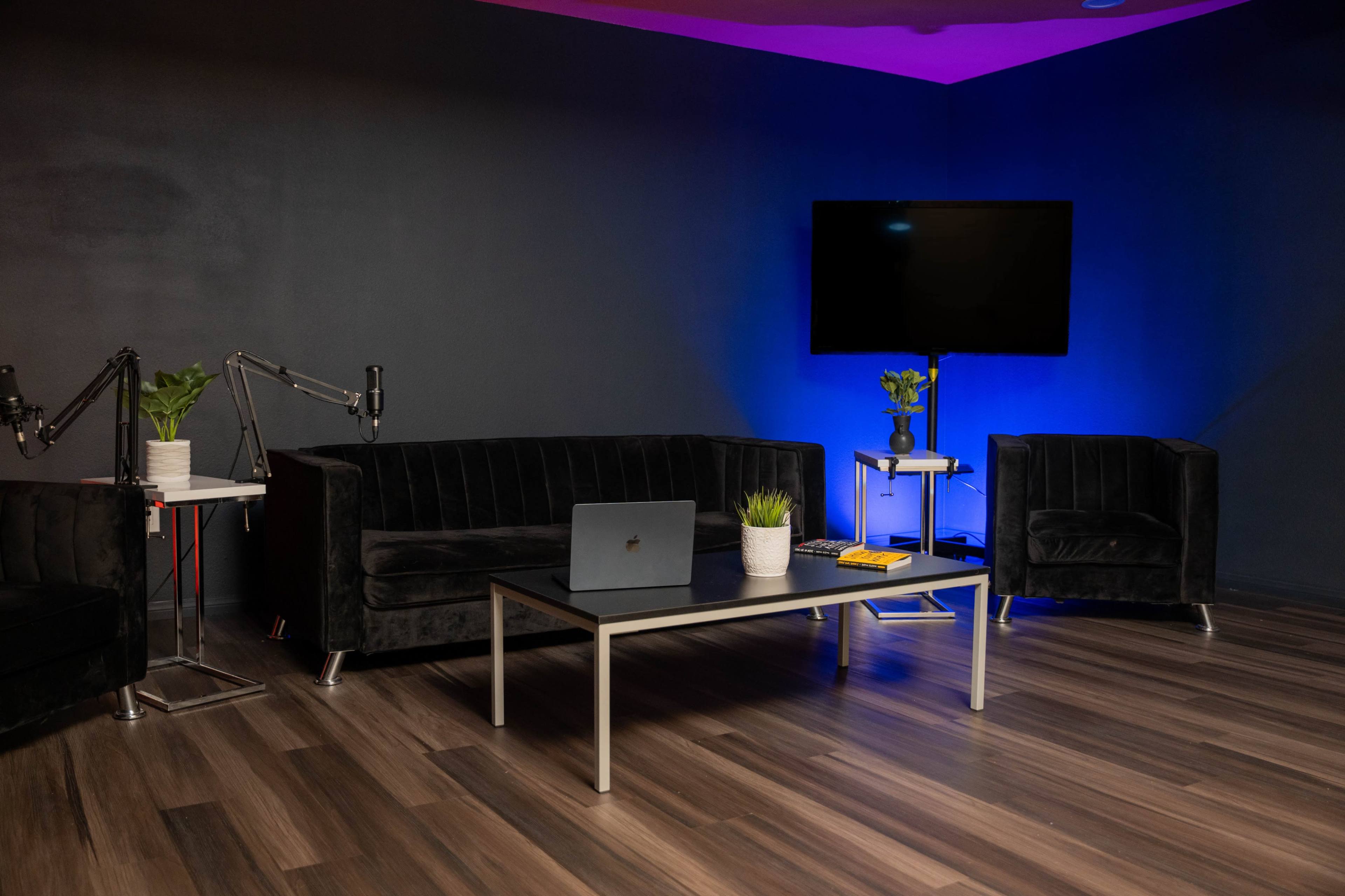 The image shows a modern living space with black furniture, including a sofa and two armchairs, a coffee table, and a large wall-mounted television, all set against a dark background with blue lighting.