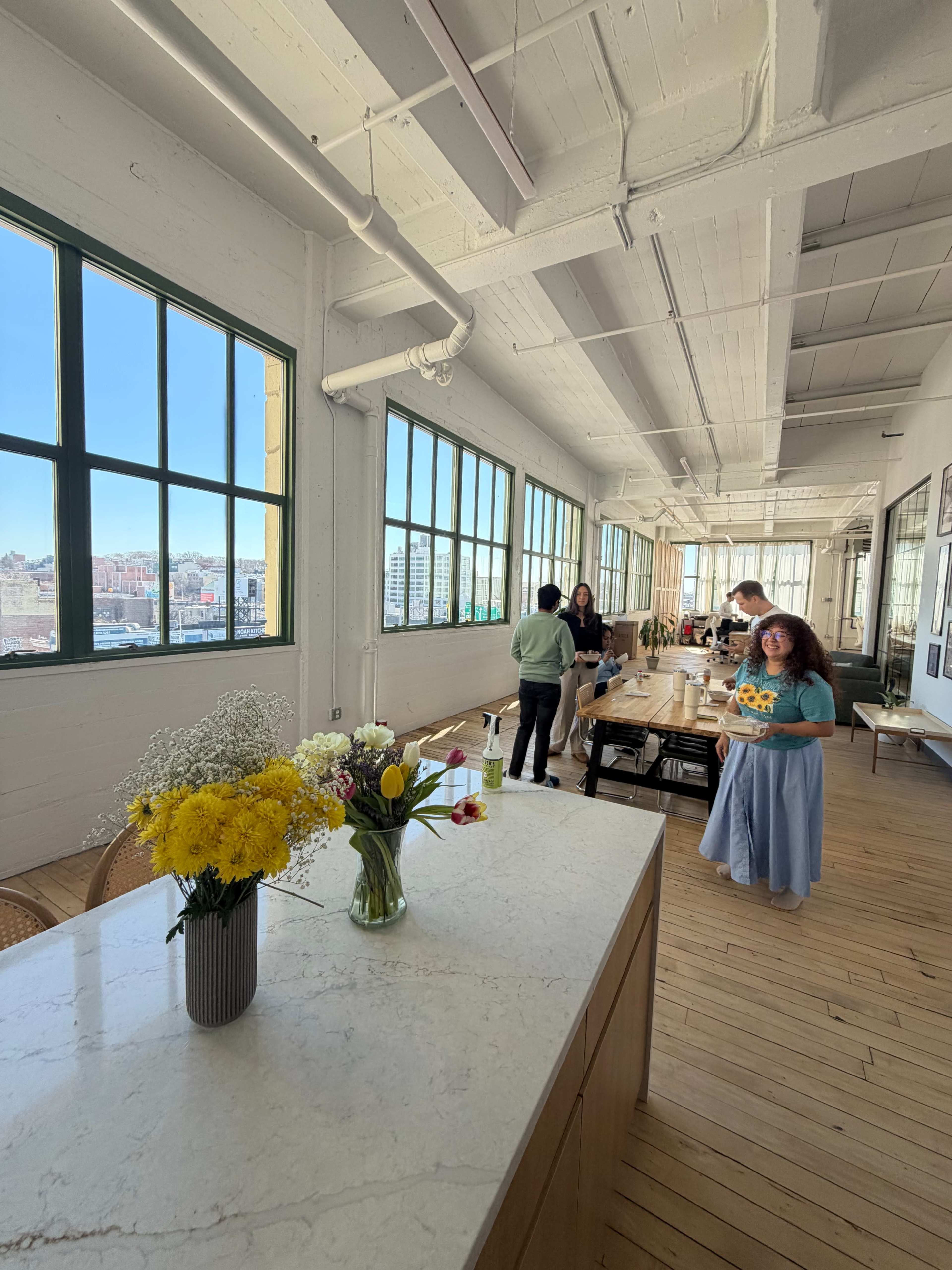 A bright, spacious room with large windows features a wooden table set for an event, complemented by flower arrangements on a marble counter.