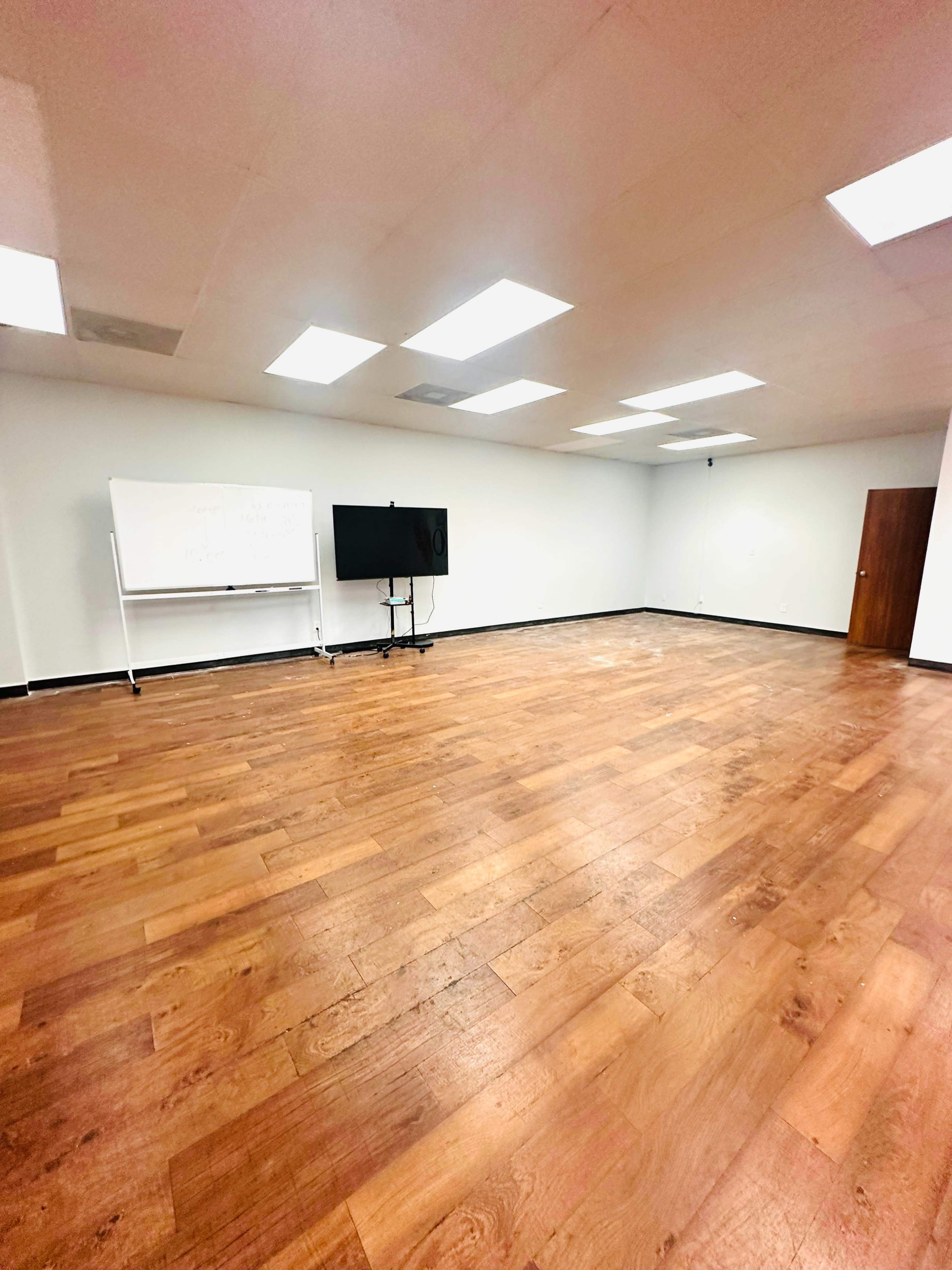The image shows a large, empty room with wooden flooring, a whiteboard on wheels, and a mounted television screen against a plain white wall.