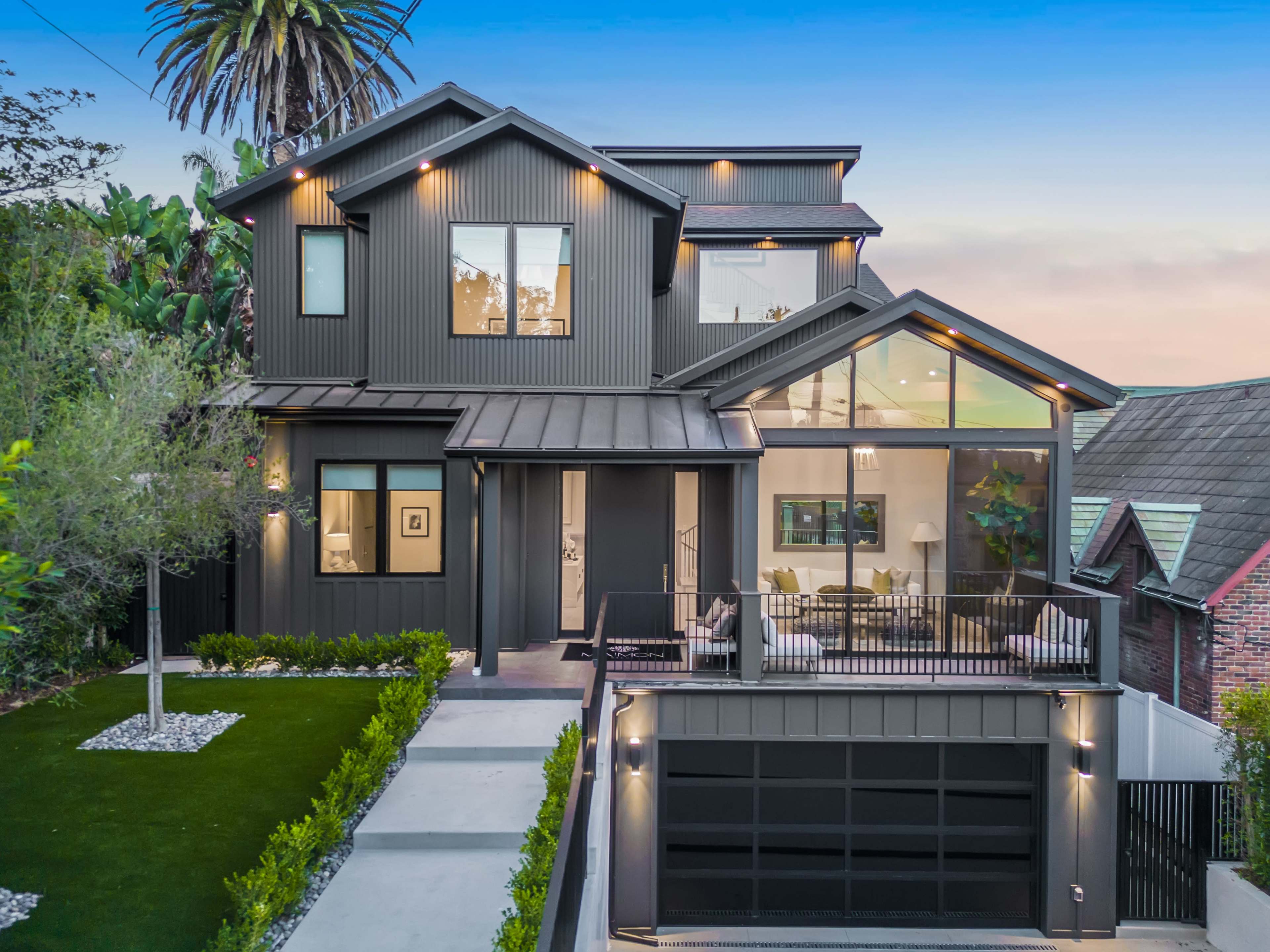 The image shows a modern two-story house with a dark exterior, large windows, and an attached garage, surrounded by greenery and a driveway.