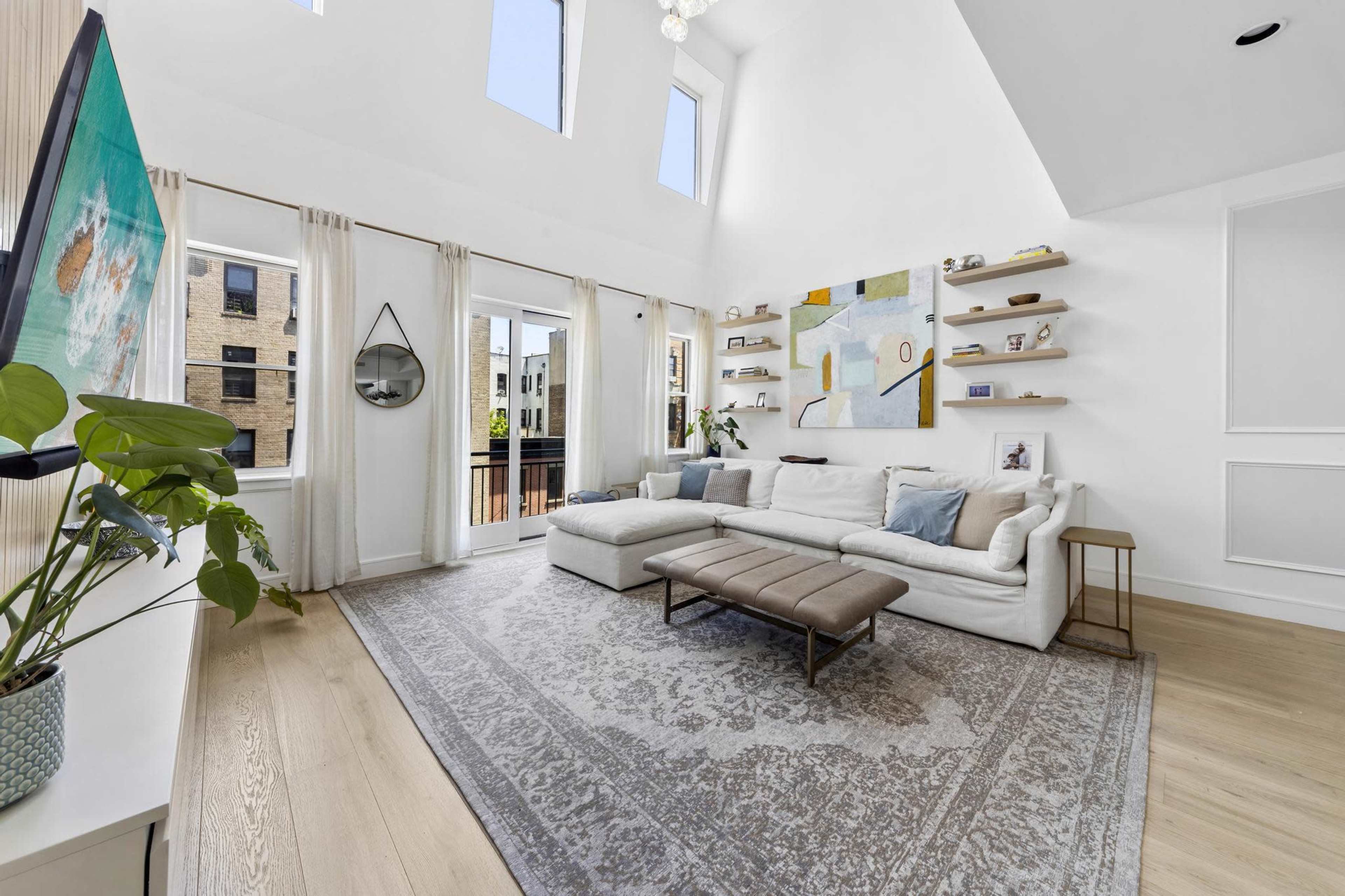 A spacious living room with high ceilings, a large sectional sofa, and a wall-mounted television, complemented by natural light streaming through several windows.
