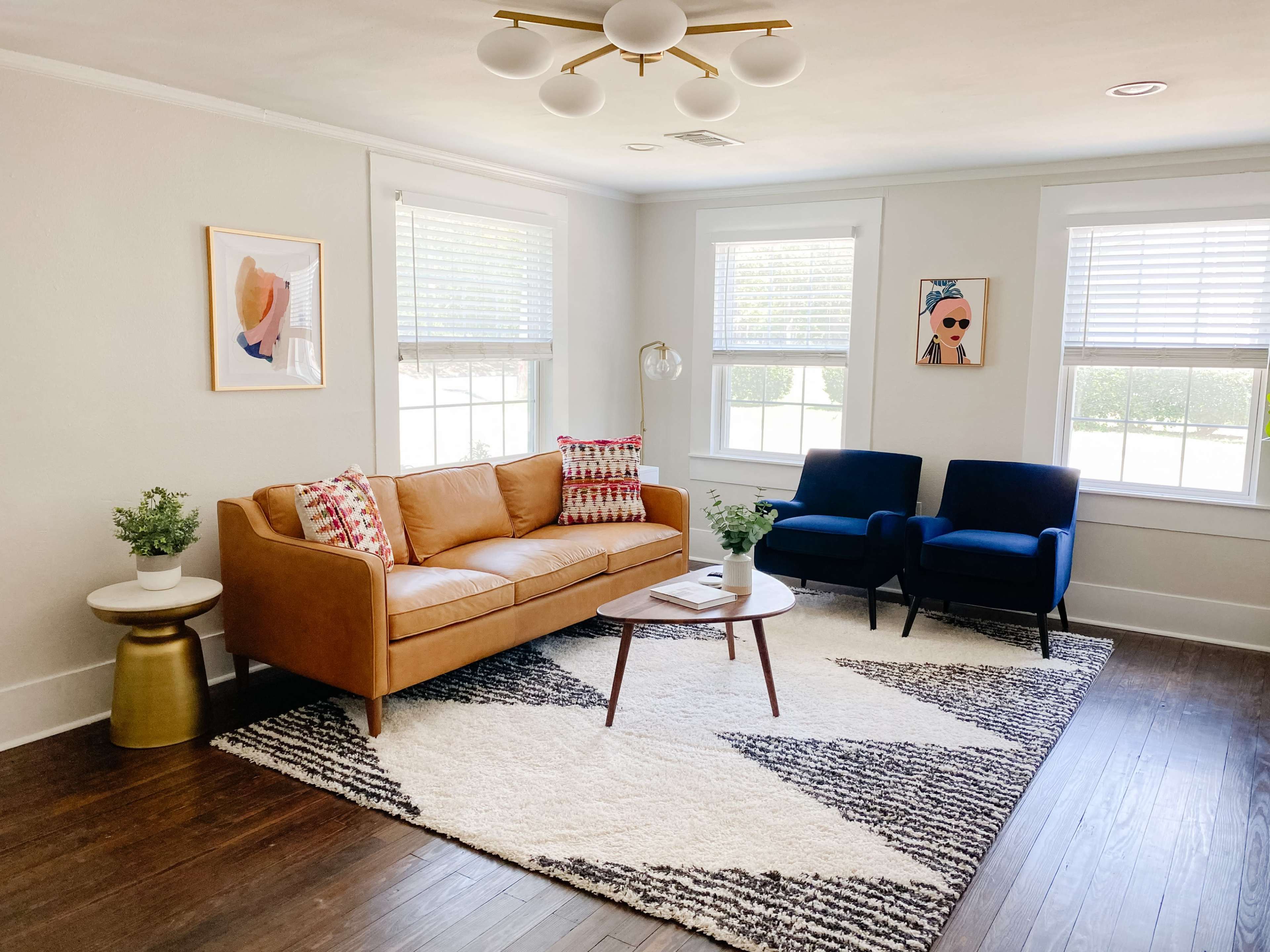 The living room features a tan leather sofa, two blue chairs, a coffee table on a patterned rug, and artwork on the walls.