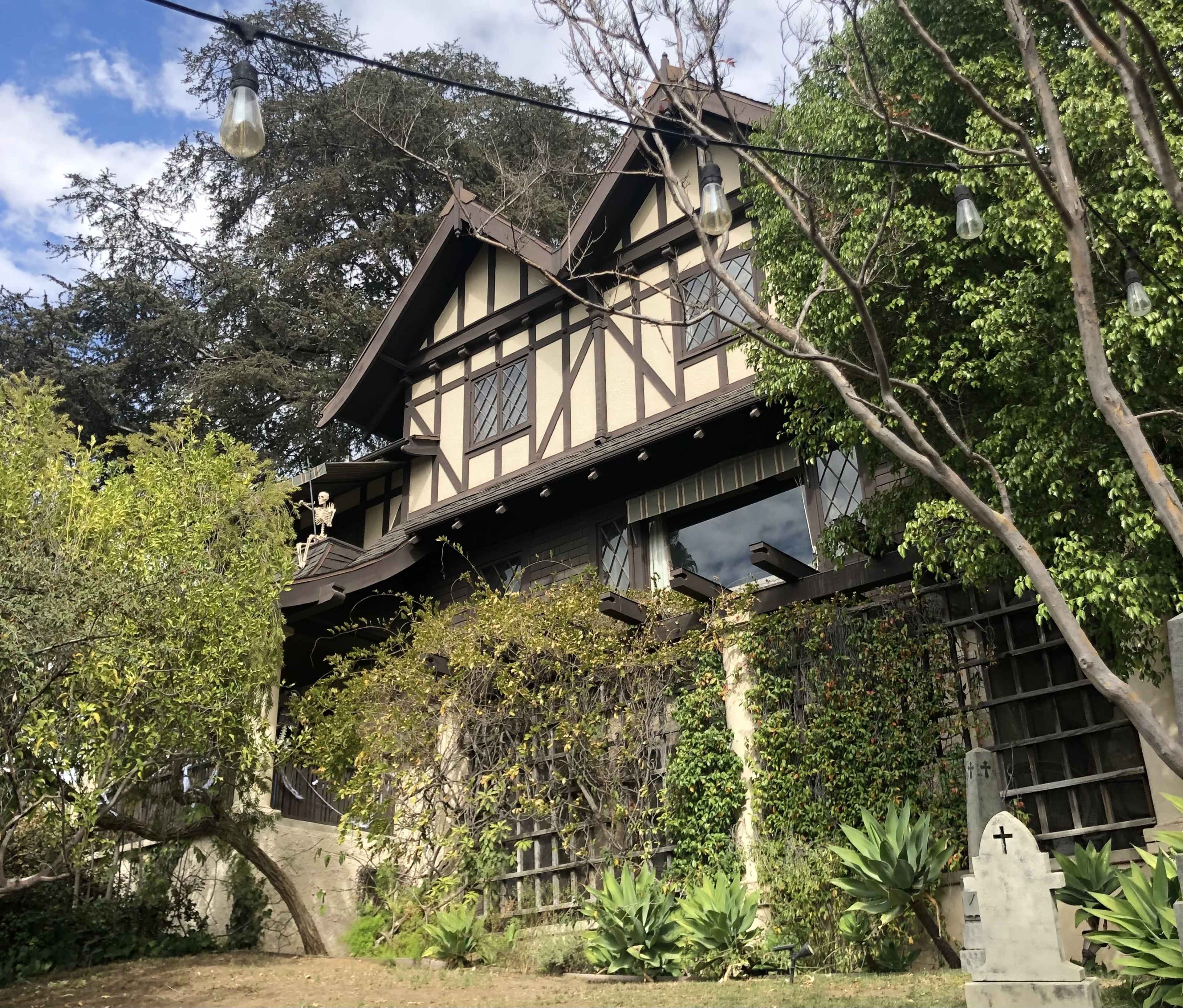 A large Tudor-style house with a stone exterior and a sloping roof is surrounded by greenery and decorative plants.