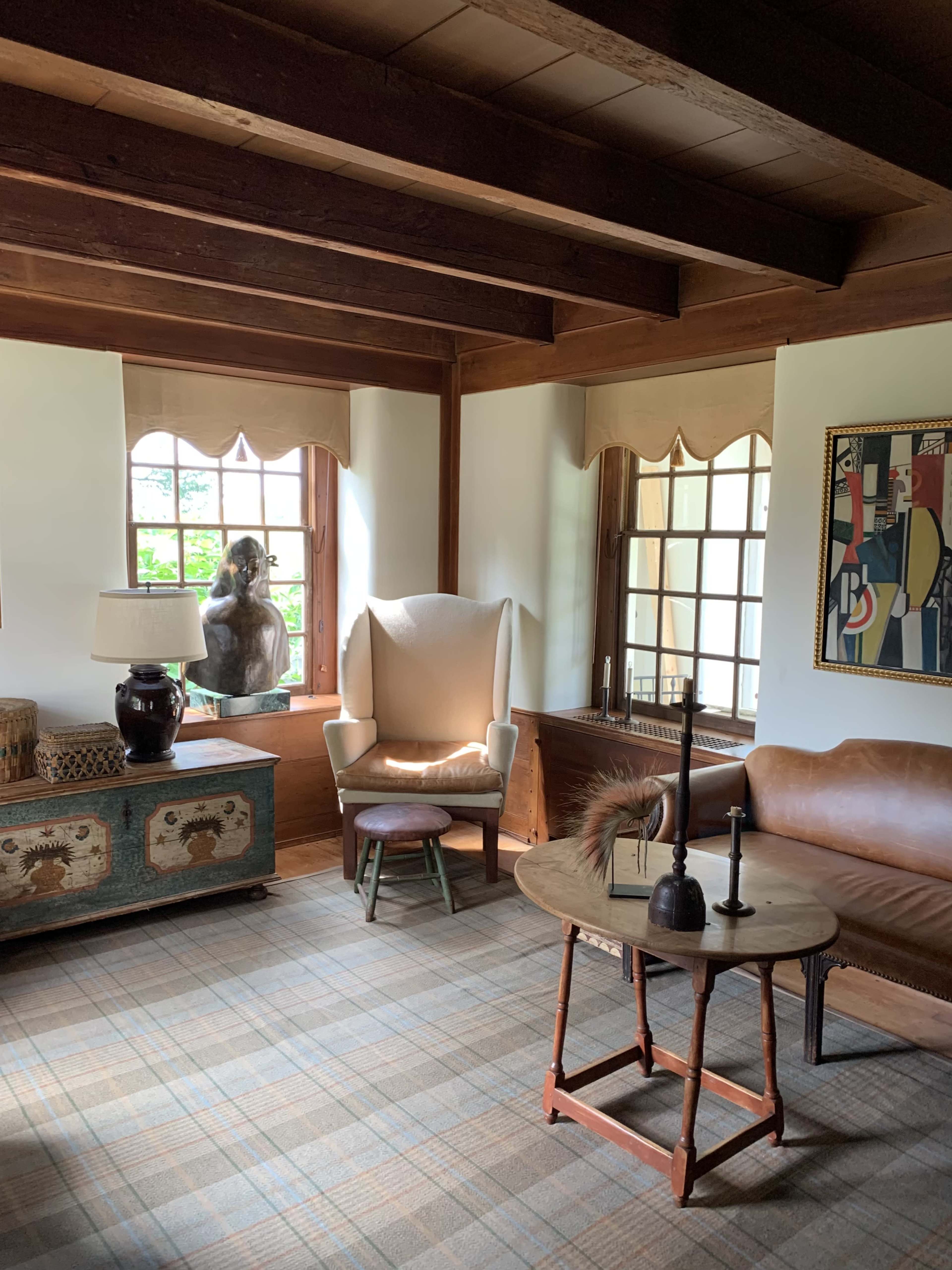The image shows a cozy living room with wooden beams, two windows, a light-colored armchair, a leather sofa, a round coffee table, and decorative items on the table and shelves.