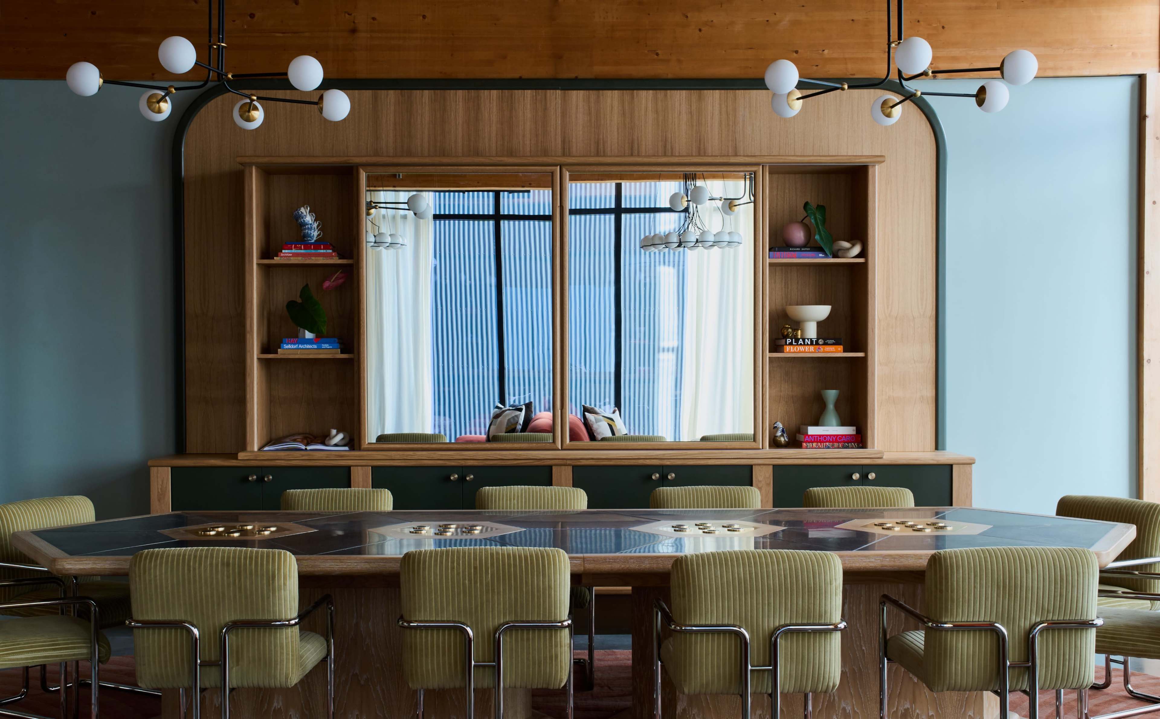 A modern dining room features a wooden table surrounded by upholstered chairs, with a wall-mounted shelving unit reflecting a large window in the background.