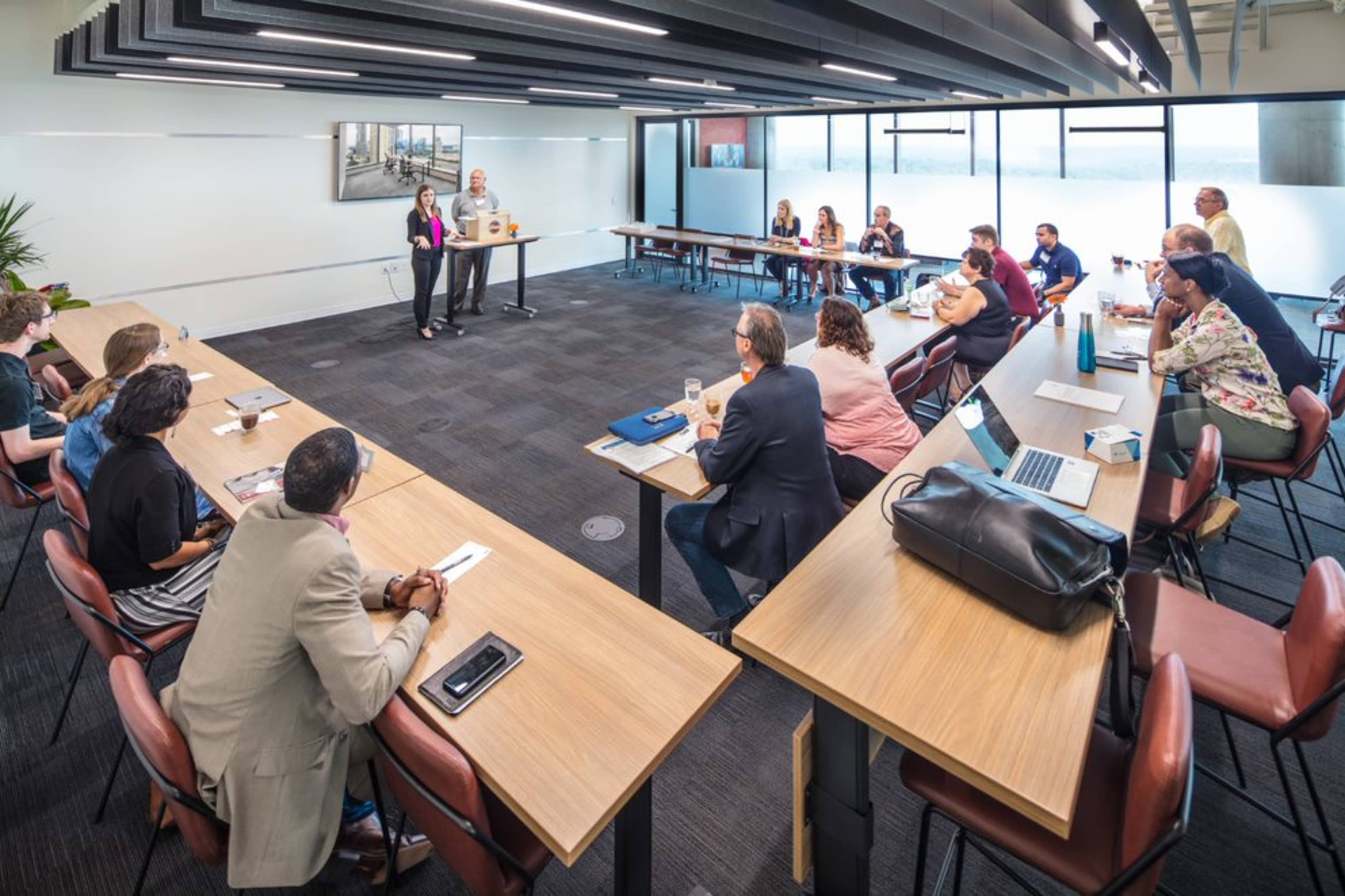 A group of people is seated at long tables in a conference room, while two speakers present in front of a large window.