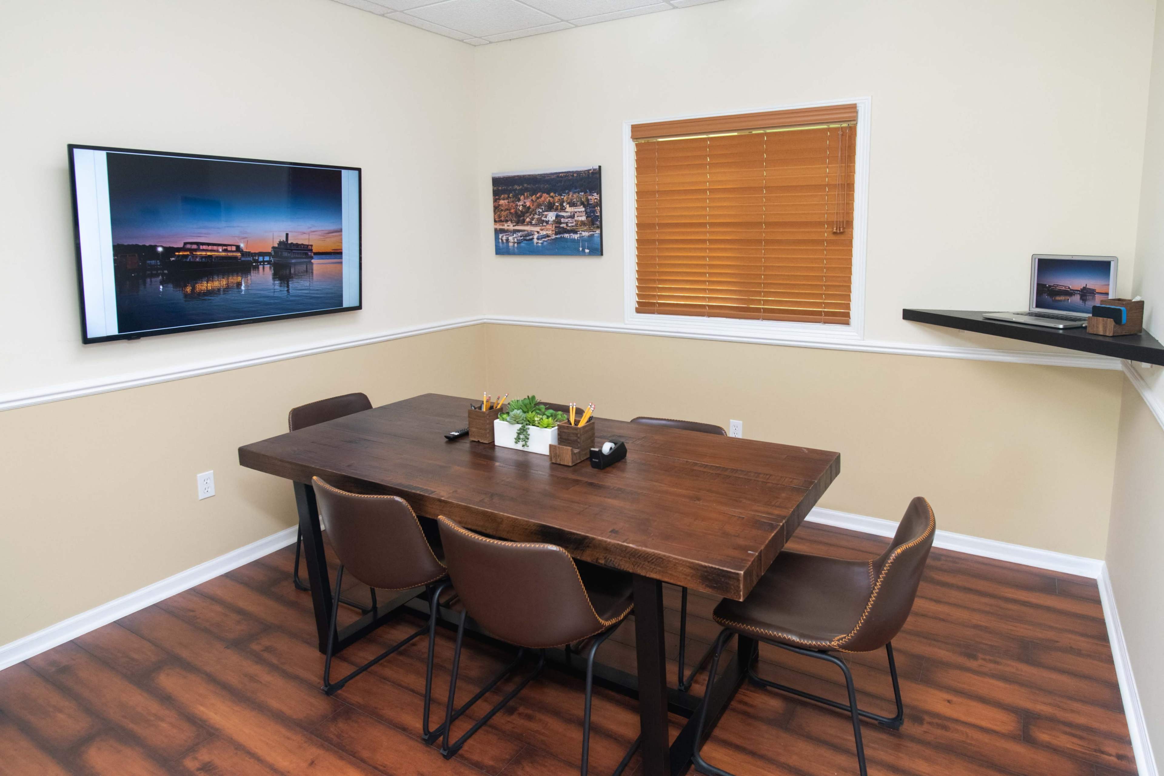 A conference room features a wooden table surrounded by chairs, with a television mounted on the wall and artwork displayed near a window with blinds.