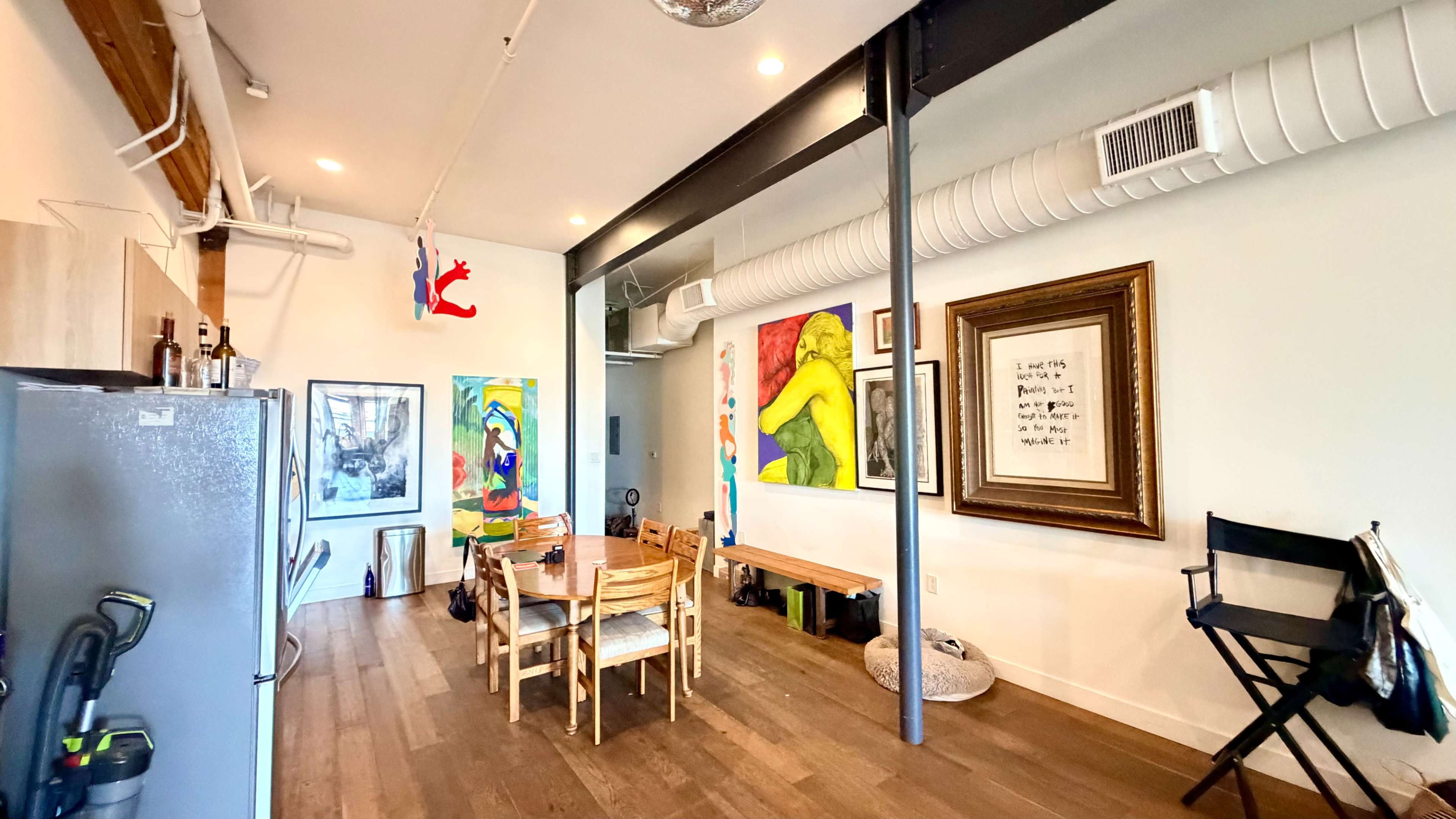 The image shows a modern open-concept living space featuring a dining area with a round table surrounded by chairs, eclectic artwork on the walls, and a kitchen area with a refrigerator and minimalist decor.