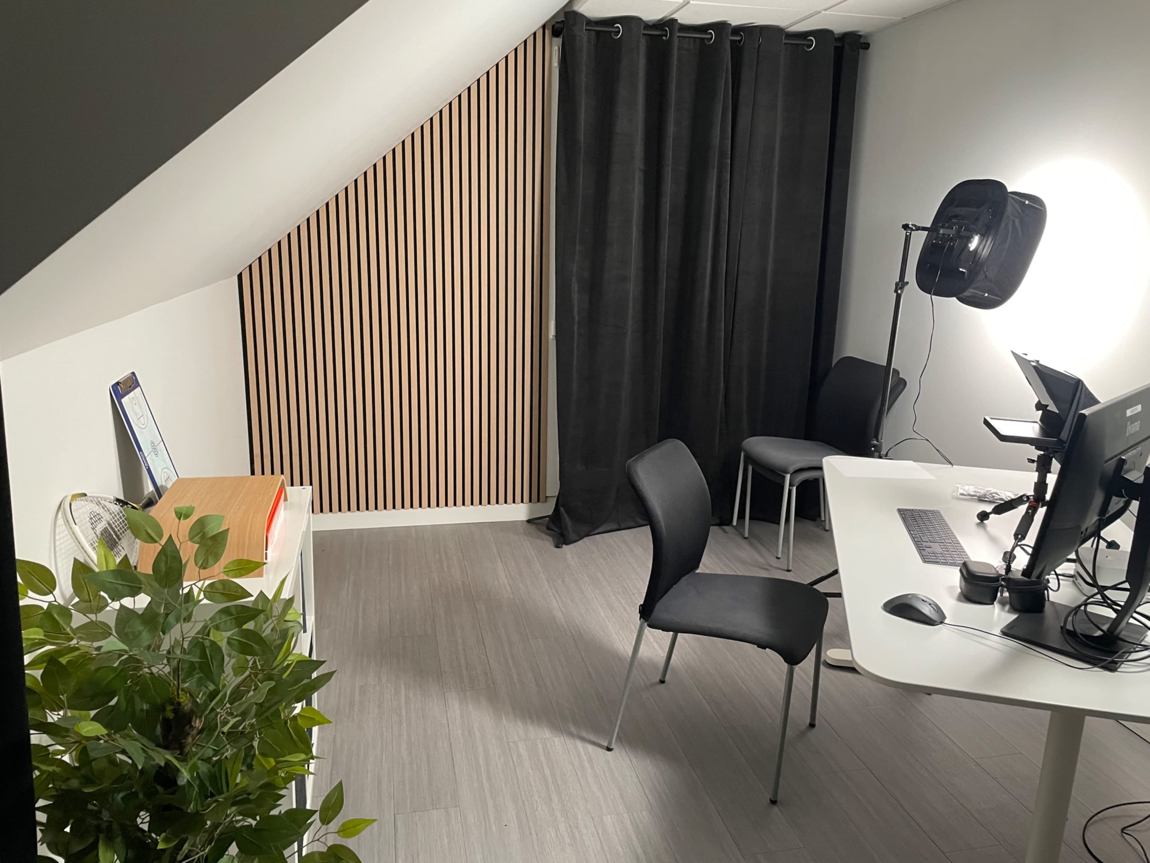 The image shows a small office space with a desk, a computer monitor, two black chairs, and a houseplant, set against a backdrop of curtains and a wooden panel wall.