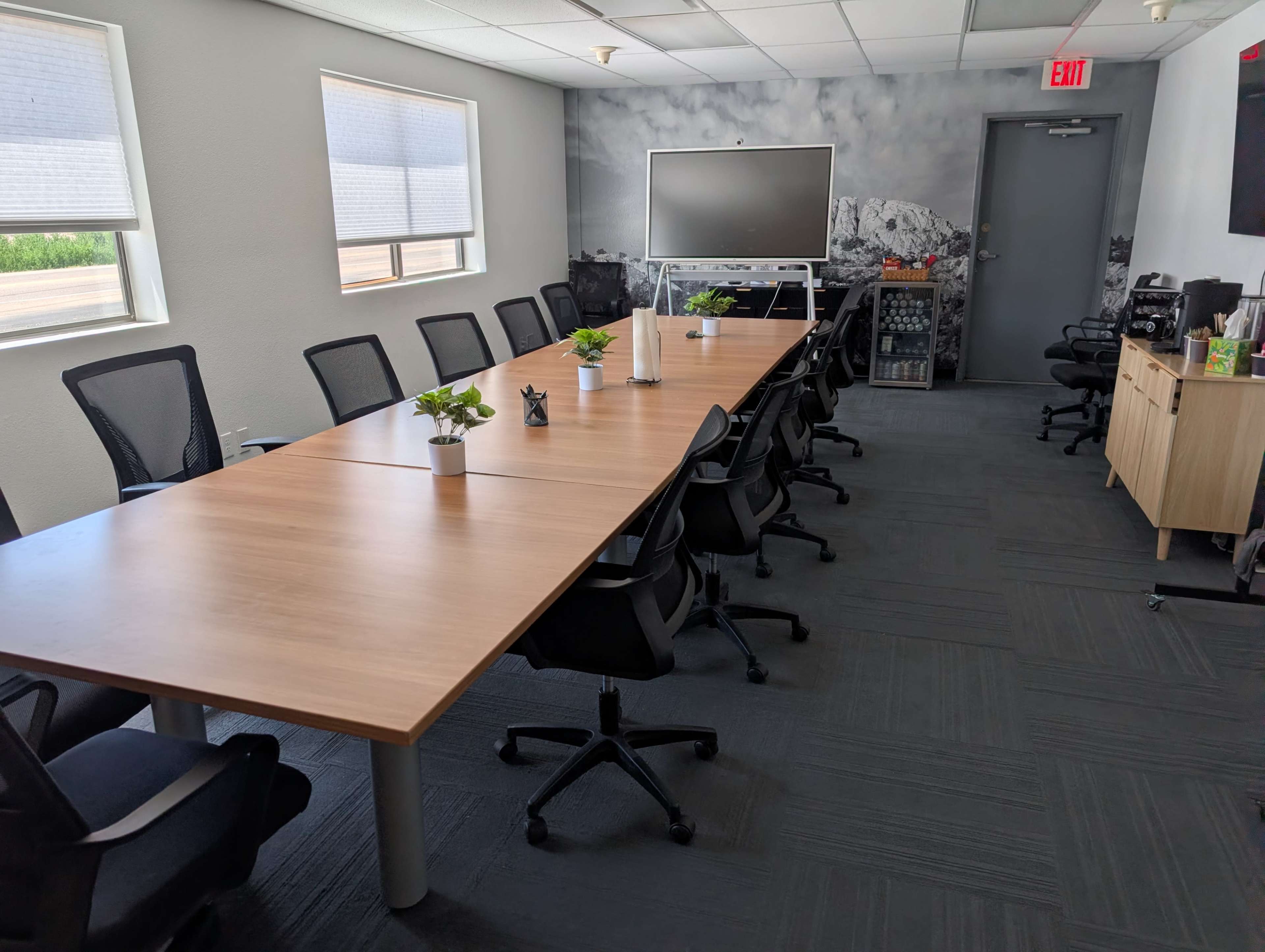 The image shows a spacious conference room with a long wooden table, black office chairs, and a large screen at one end.