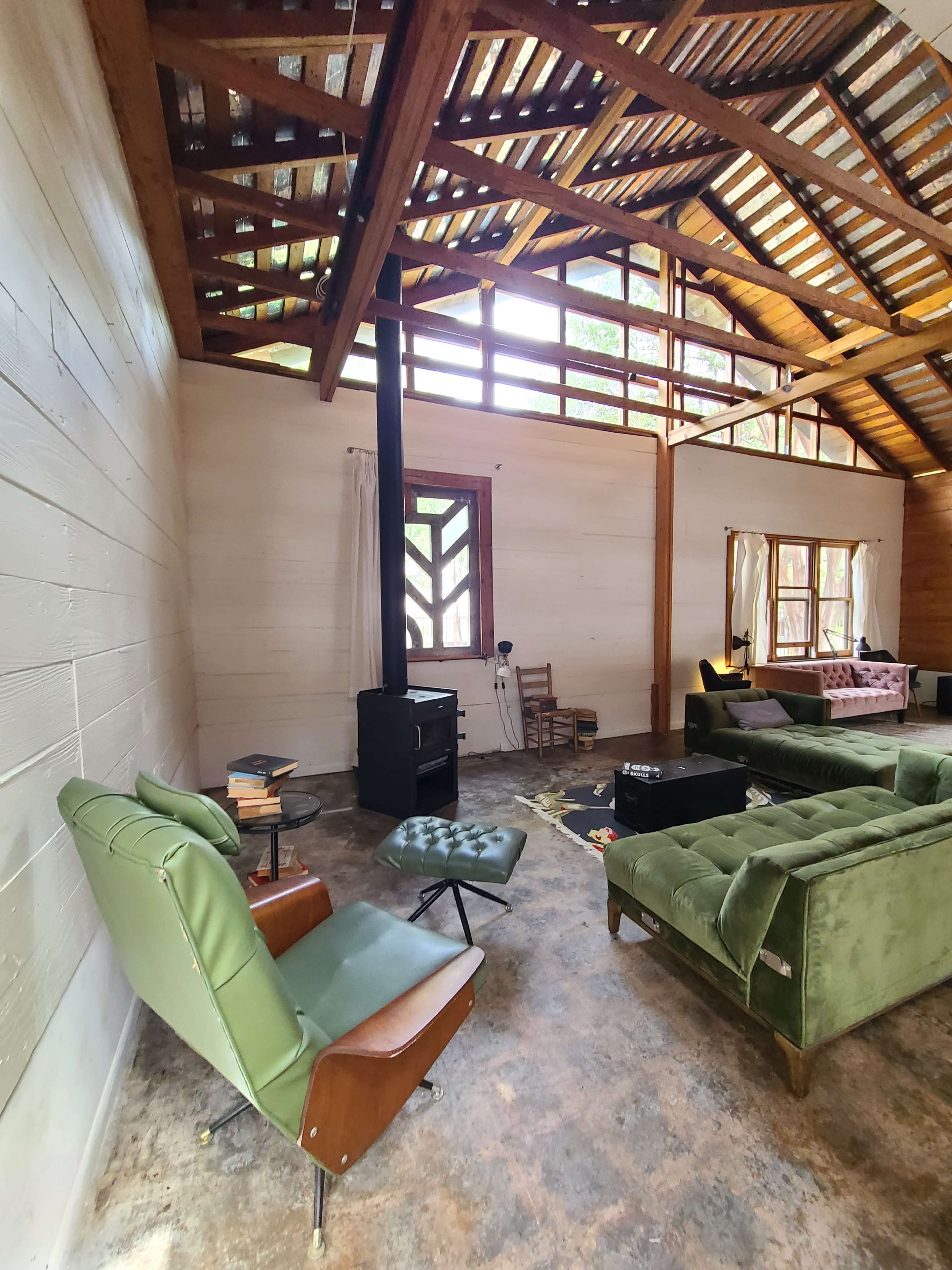 A cozy living room with green vintage chairs, a wood stove, and large windows allowing natural light to enter.