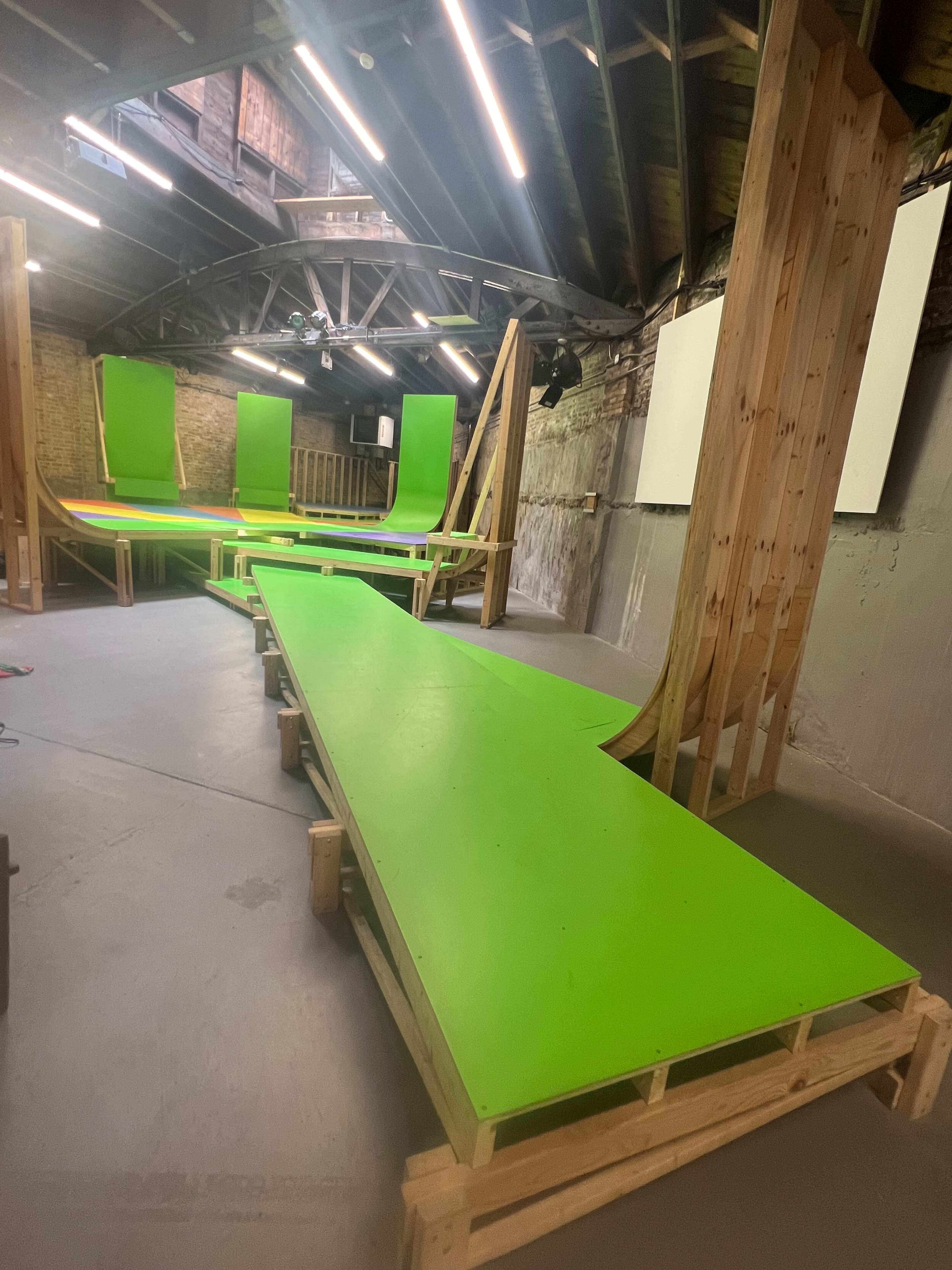 The image shows a skateboard ramp setup made of wooden structures and vibrant green panels inside a warehouse with exposed brick walls.