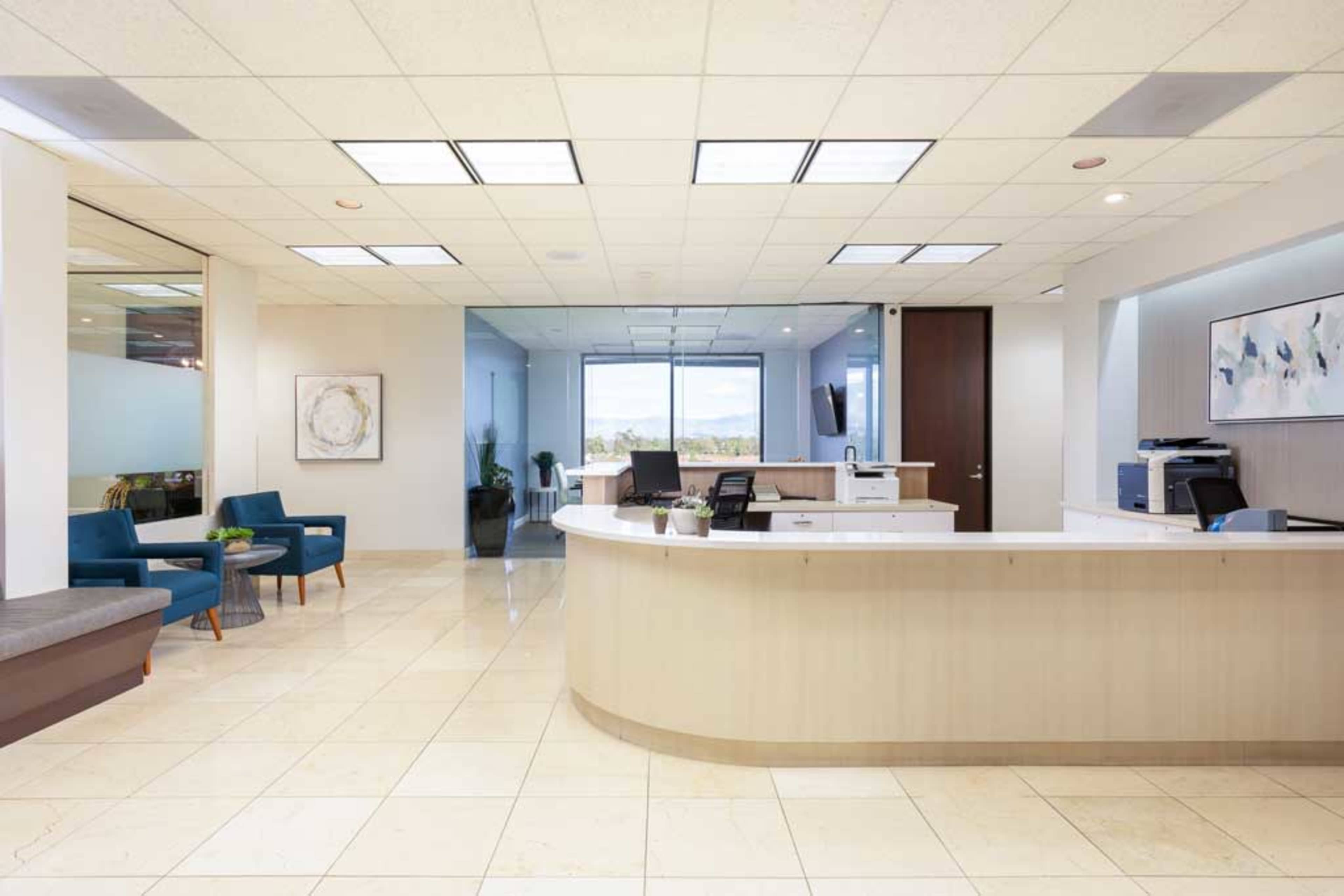 A modern reception area features a curved front desk, seating areas with blue chairs, and large windows allowing natural light.