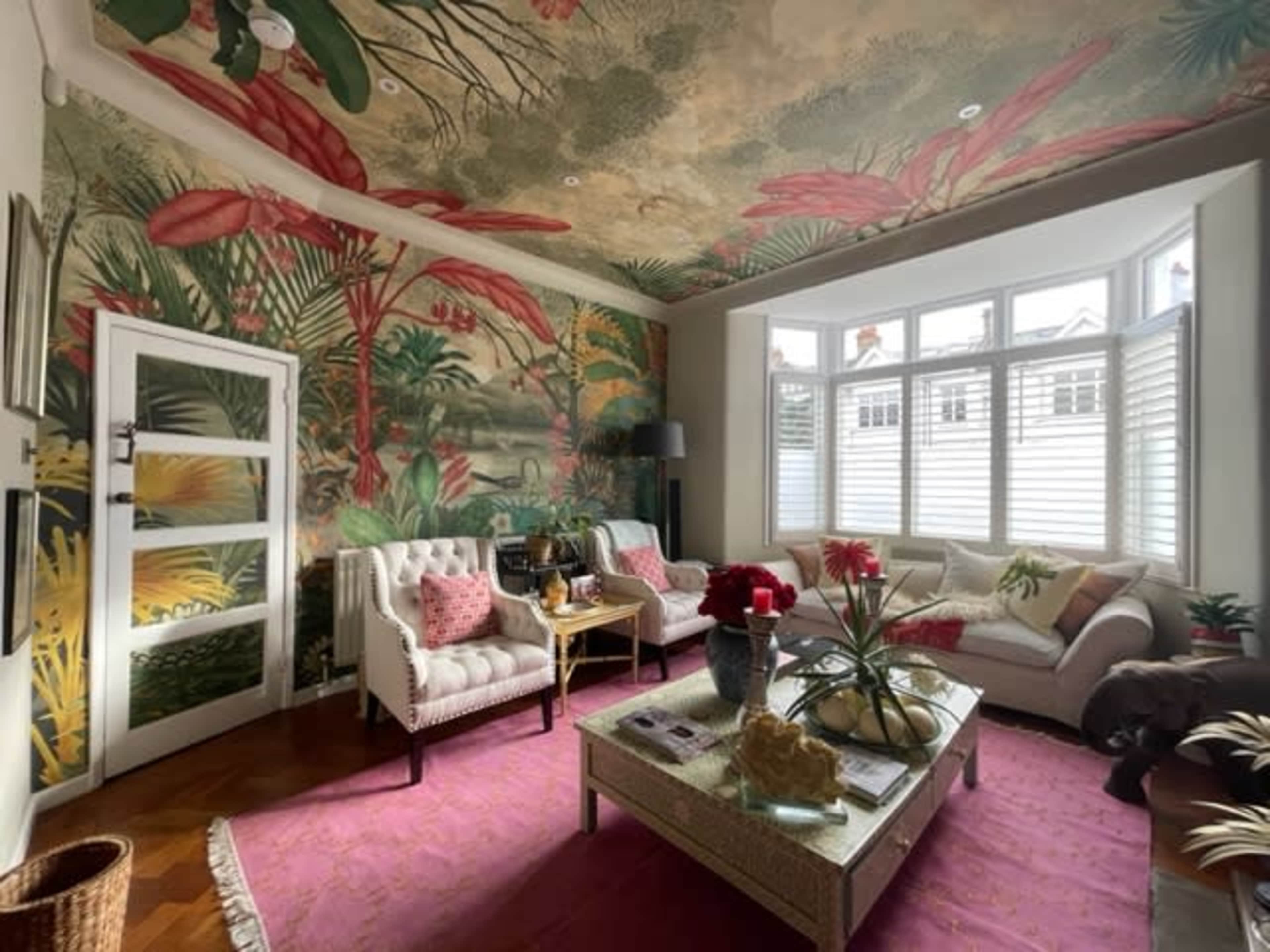 A living room features tropical-themed wallpaper, oversized plants in the mural, modern furniture, and large windows with white shutters.