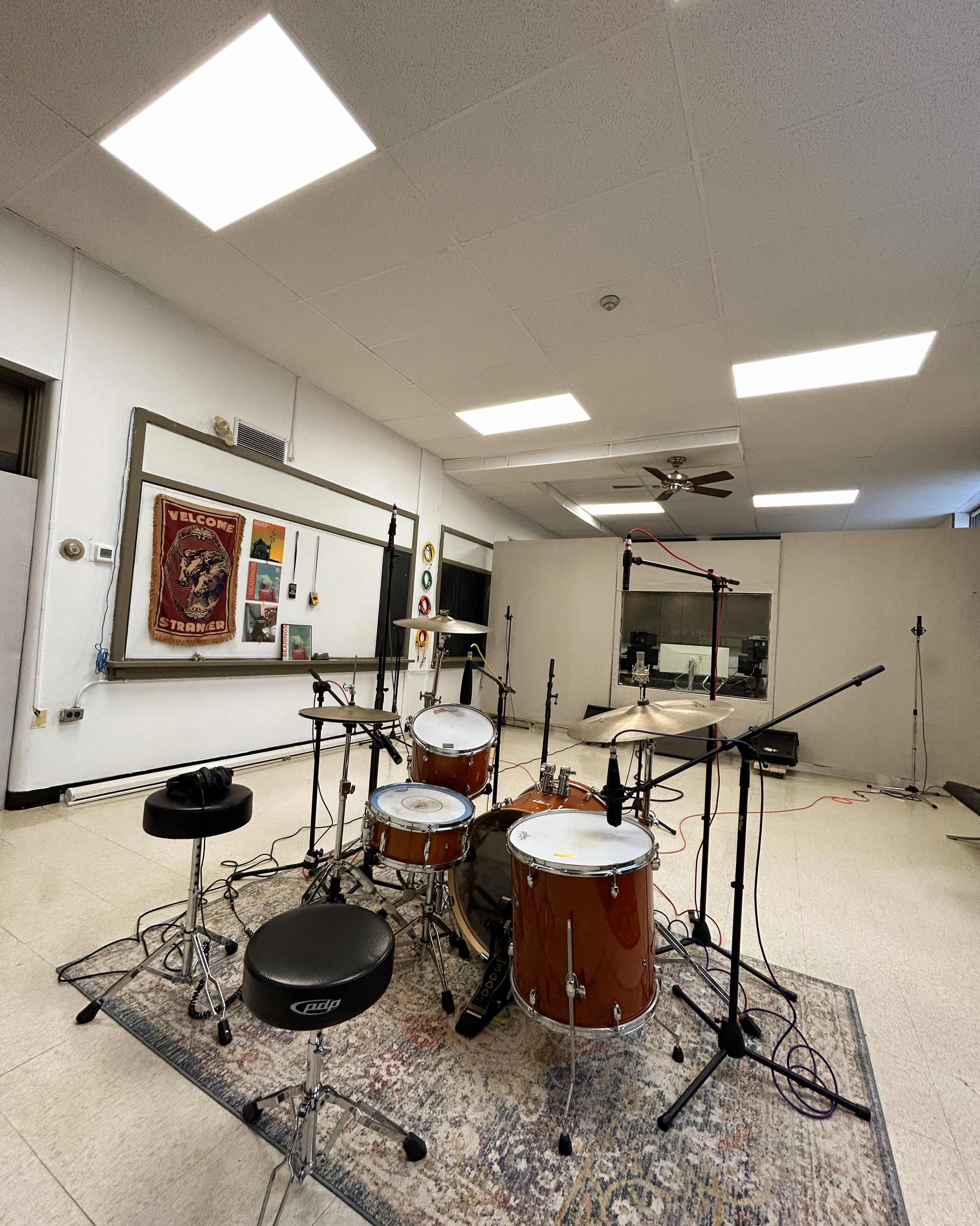 A drum set is positioned in a well-lit recording studio with microphones set up around it.